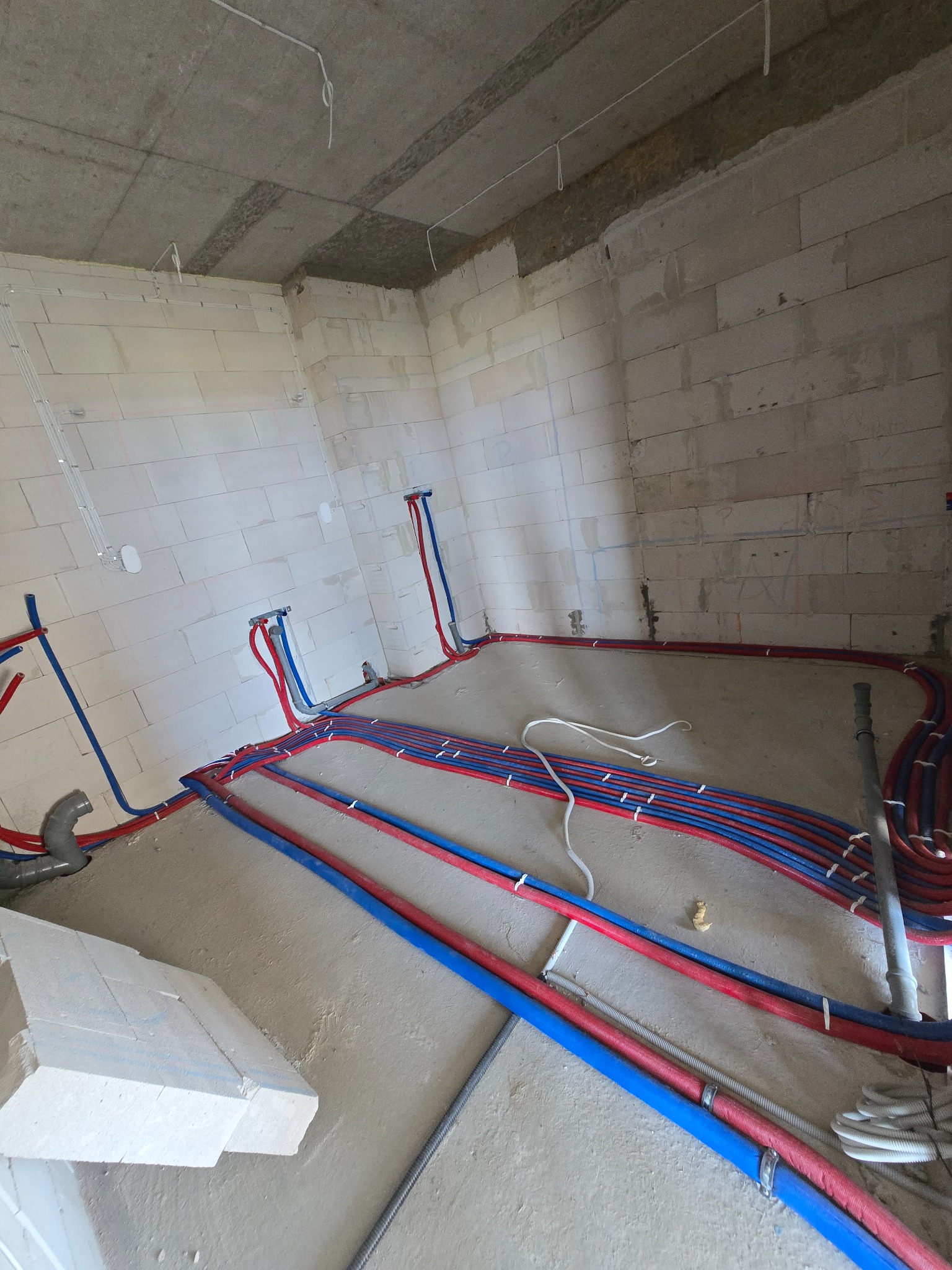 Surowe wnętrze w trakcie prac hydraulicznych: czerwono-niebieskie rury grzewcze ułożone na podłodze, wyprowadzone na ścianach z betonu komórkowego.