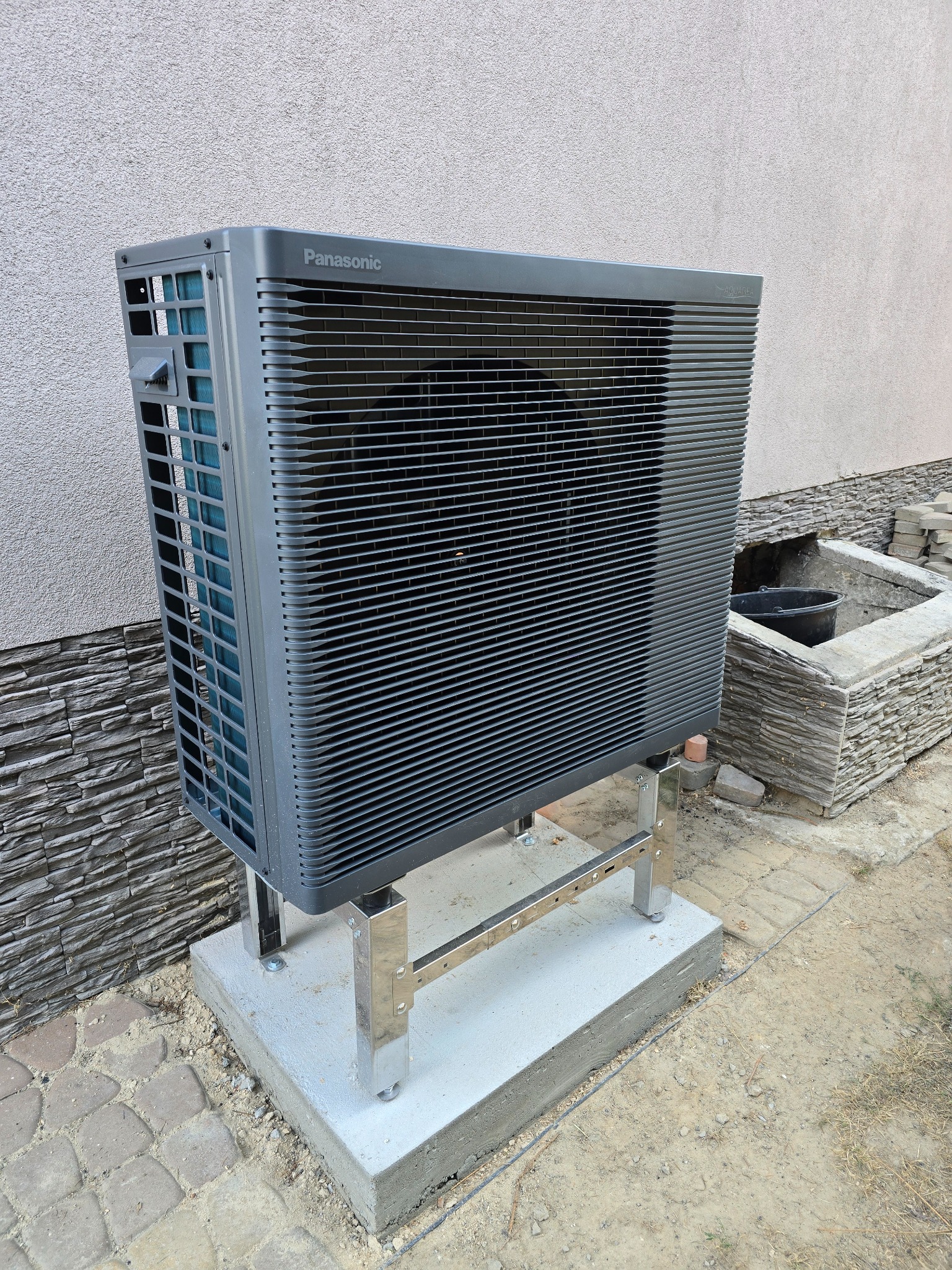 Szara pompa ciepła Panasonic Aquarea na betonowej podstawie, zamontowana przy ścianie budynku. Widoczna metalowa konstrukcja wsporcza i fragment otoczenia.