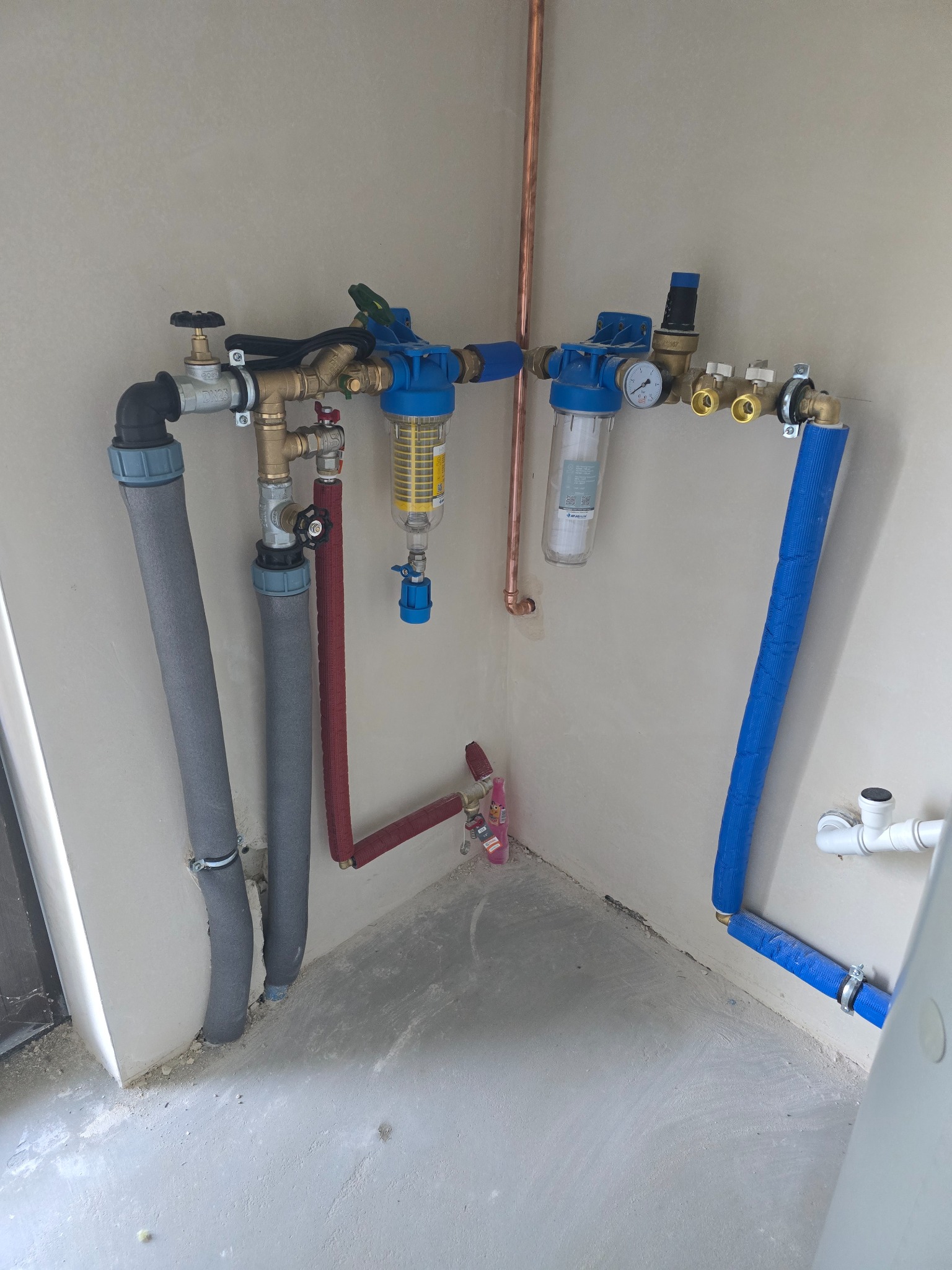 Kompleksowa instalacja wodna z filtrami, zaworami i rurami w izolacji, wykonana w narożniku pomieszczenia. Widoczne elementy miedziane i plastikowe.