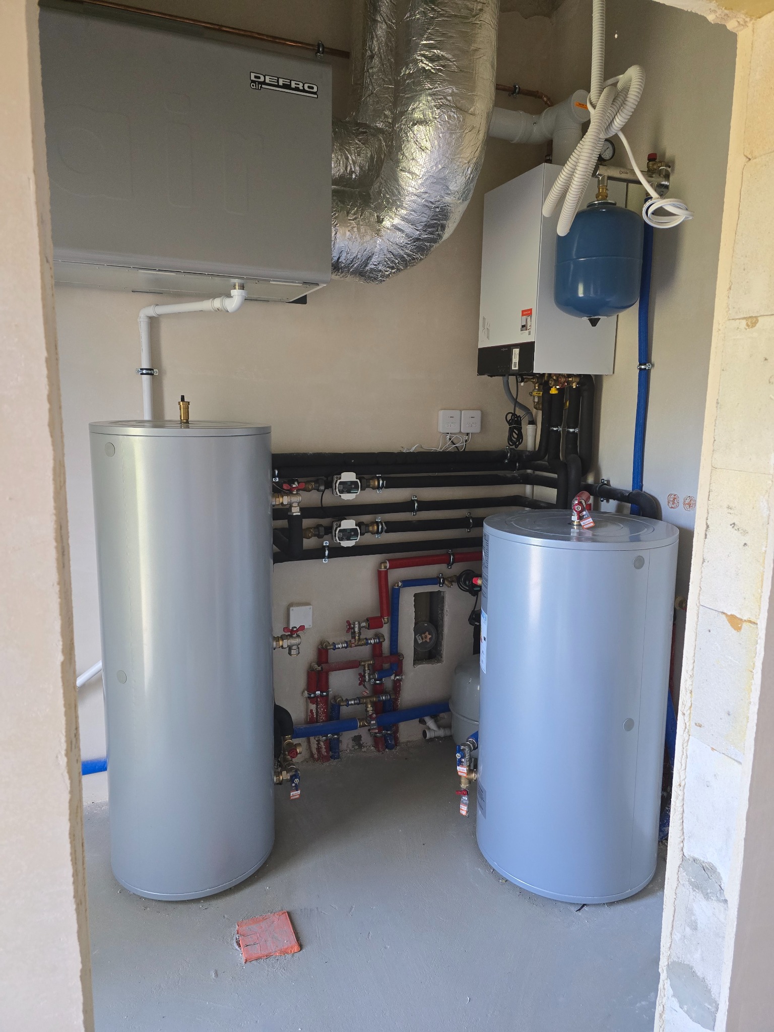 Zestaw urządzeń grzewczych: dwa zbiorniki, piec gazowy Viessmann z niebieskim naczyniem wzbiorczym i jednostka Defro Air, połączone rurami w kotłowni.