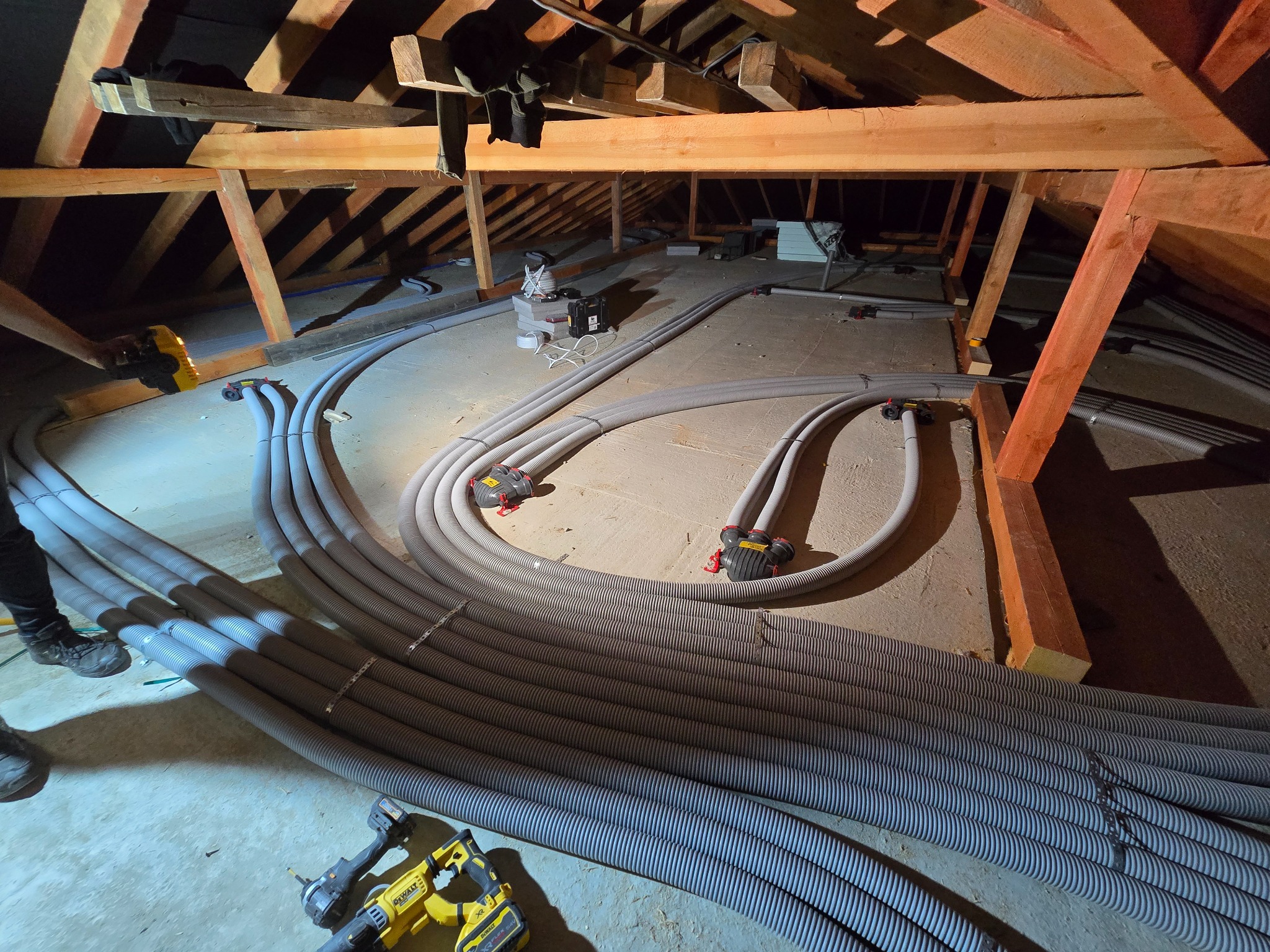 Instalacja przewodów wentylacyjnych w przestrzeni poddasza, widoczne narzędzia, drewniana konstrukcja dachu i fragment nogi instalatora.