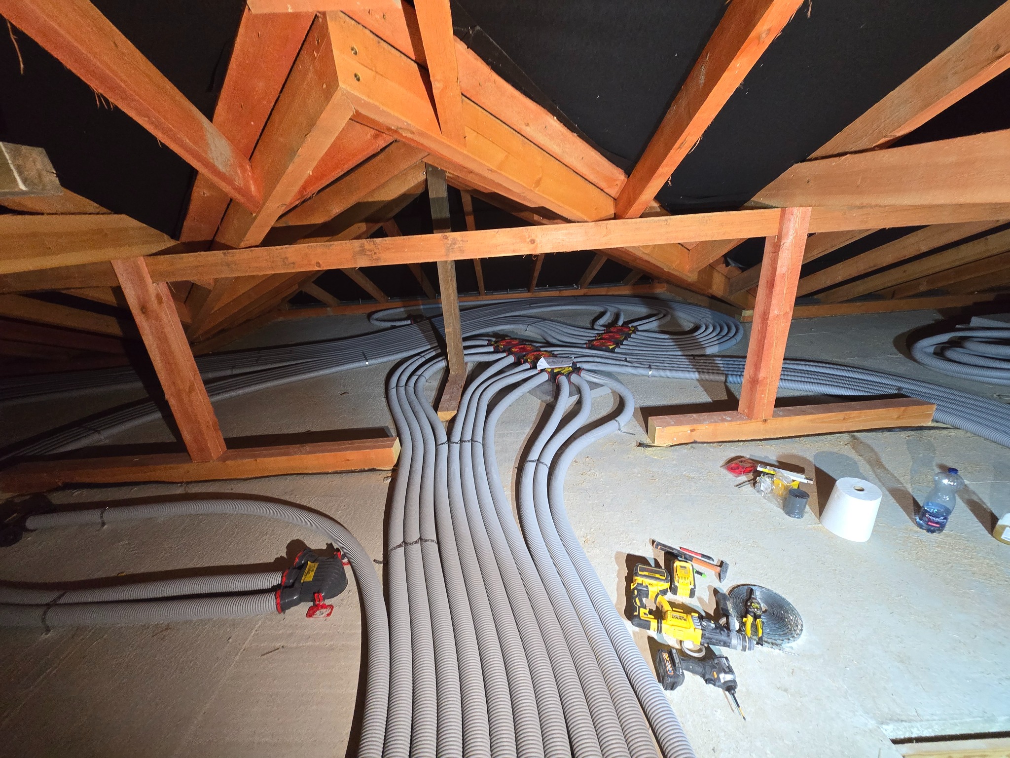 Szereg szarych rur w przestrzeni poddasza, pomiędzy drewnianymi krokwiami. Widoczne narzędzia pracy i elementy montażowe. Instalacja w trakcie realizacji.
