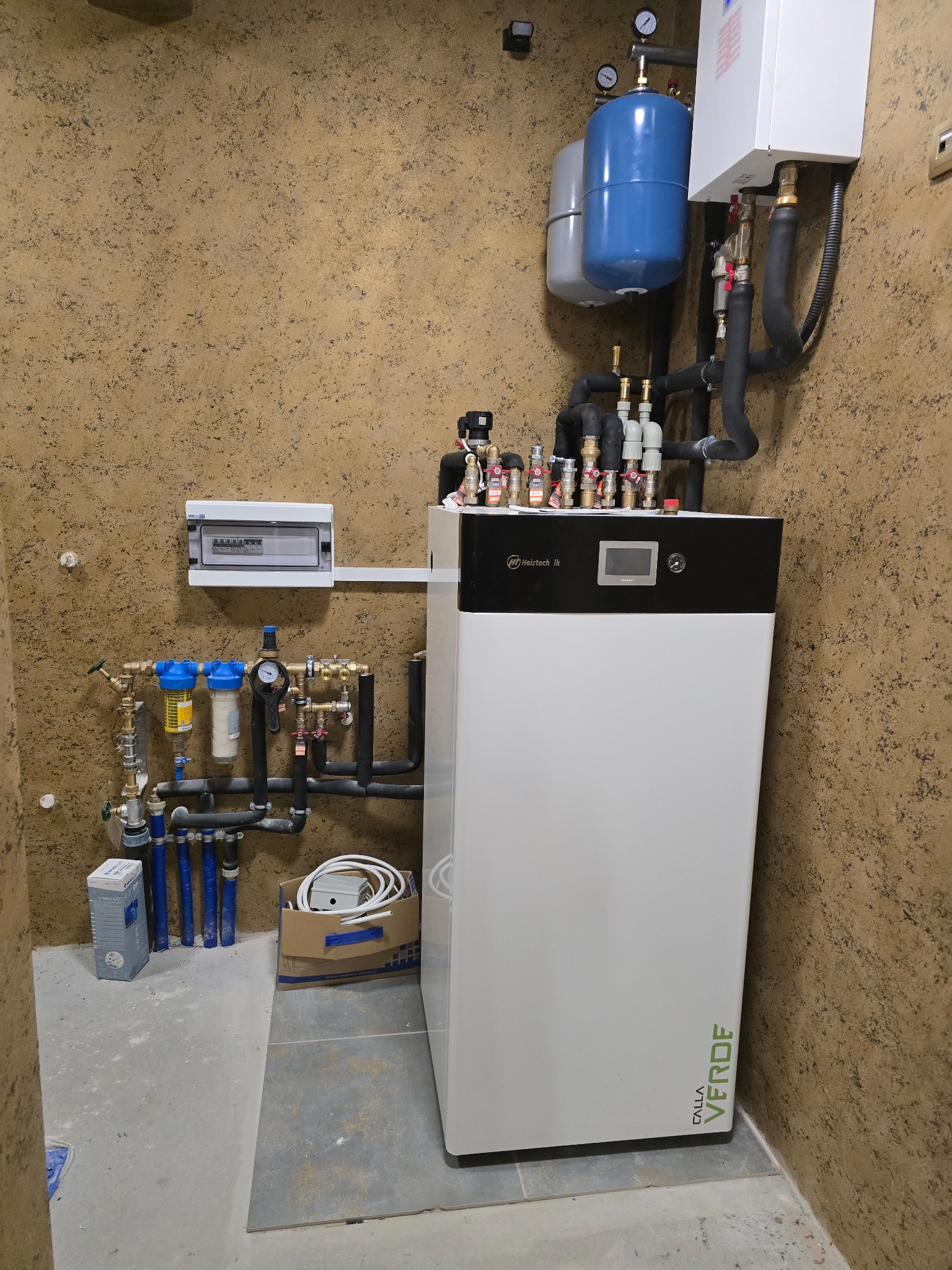 Nowoczesny, biały piec gazowy Calla Verde z podłączeniami hydraulicznymi i elektrycznymi na tle ściany o fakturze baranka. Instalacja w pomieszczeniu technicznym.