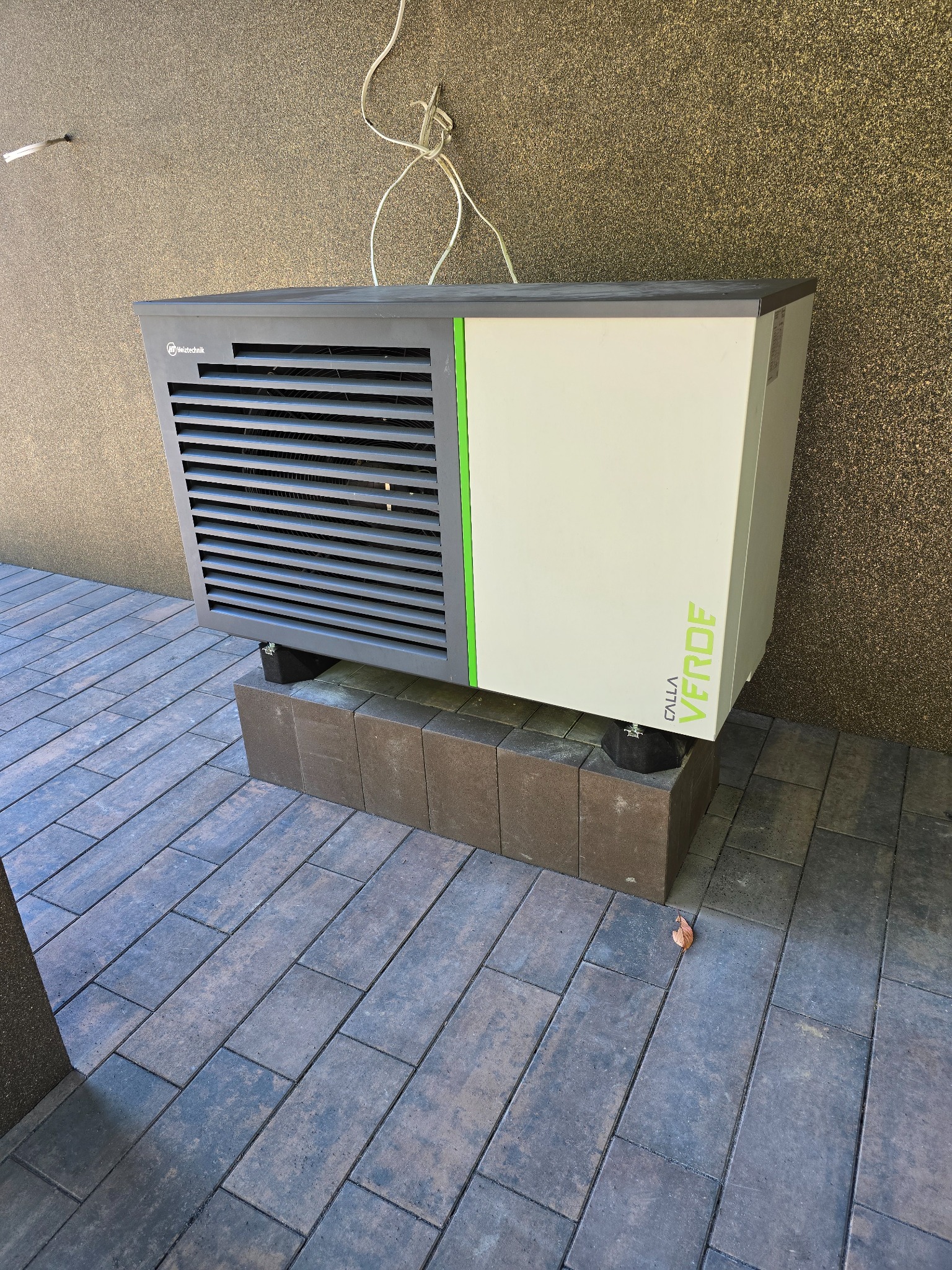 Zewnętrzna jednostka pompy ciepła Calla Verde na ceglanym cokole, z widocznymi przewodami i fakturą ściany w tle. Ujęcie z boku, na zewnątrz budynku.