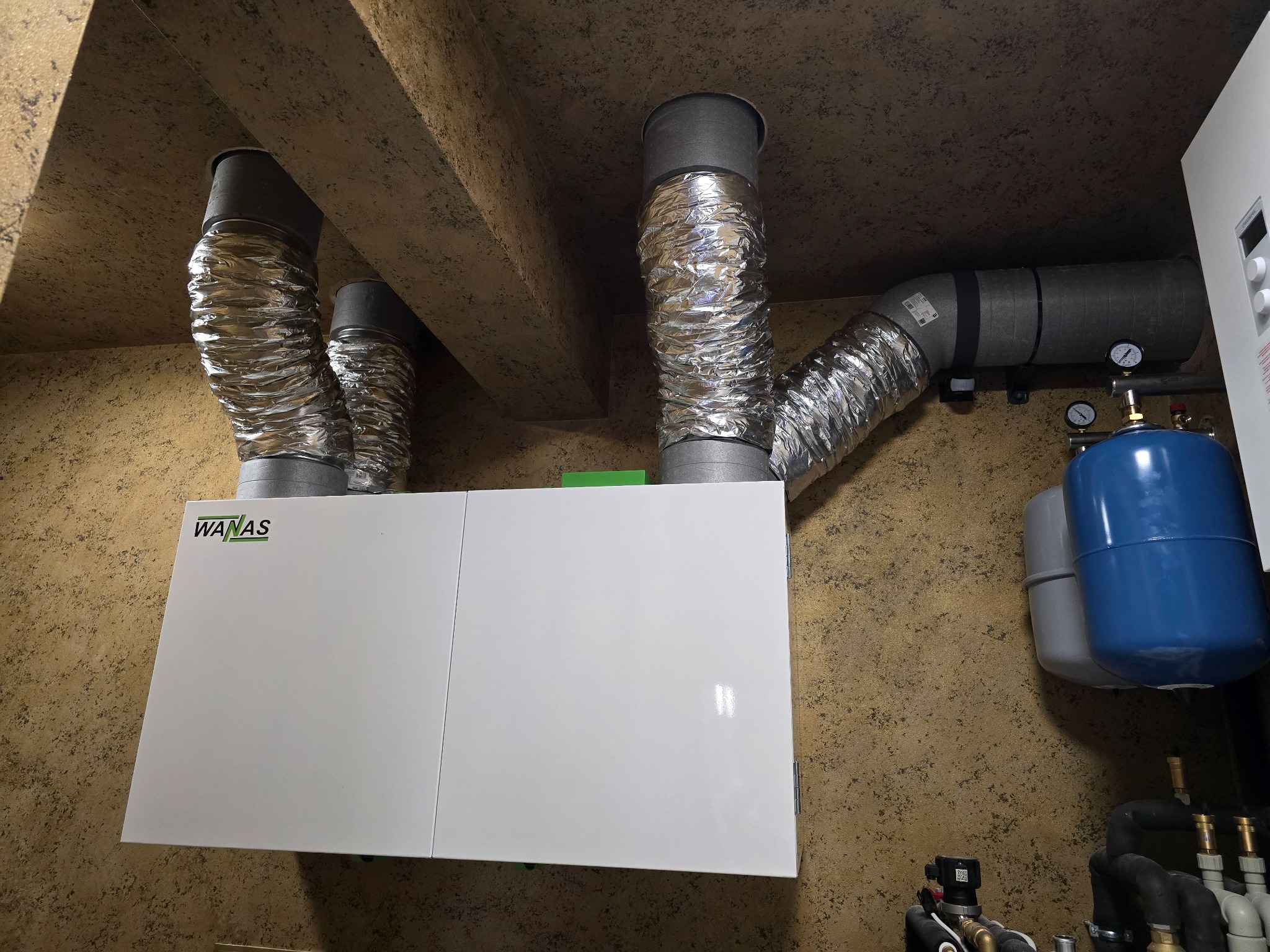 Biała centrala wentylacyjna WANAS z podłączonymi przewodami wentylacyjnymi na tle ściany o fakturze baranka. Widoczne elementy instalacji hydraulicznej.