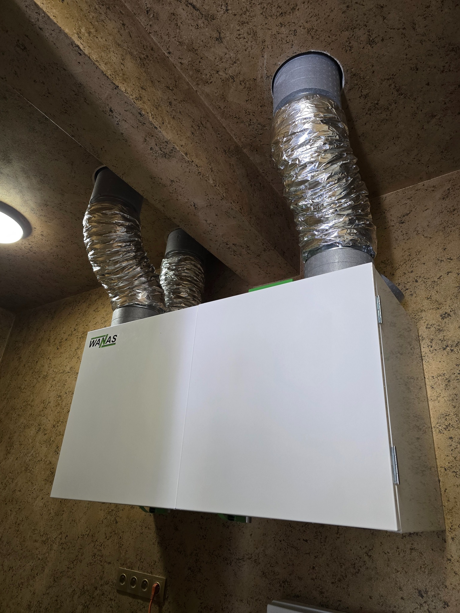 Centrala wentylacyjna WANAS z podłączonymi przewodami wentylacyjnymi, zamontowana pod sufitem w pomieszczeniu o beżowych ścianach.