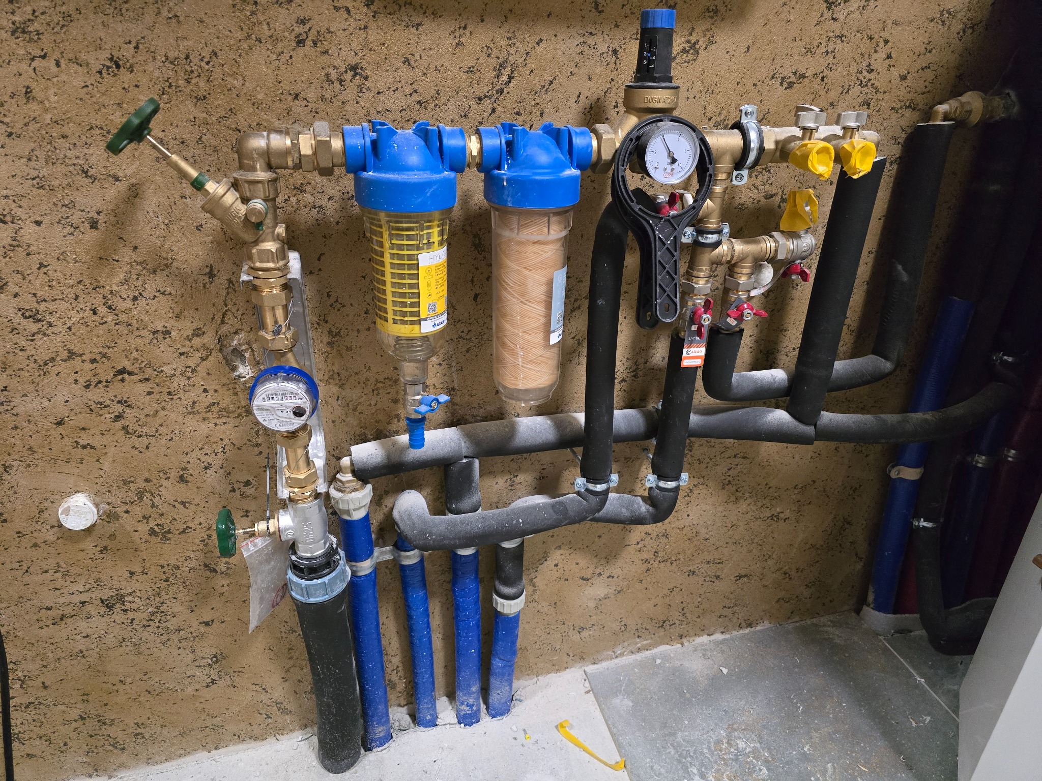 Złożona instalacja hydrauliczna z filtrami, manometrem i rurami w kolorach niebieskim i czarnym, zamontowana na beżowej ścianie. Widoczne zawory i elementy przyłączeniowe.