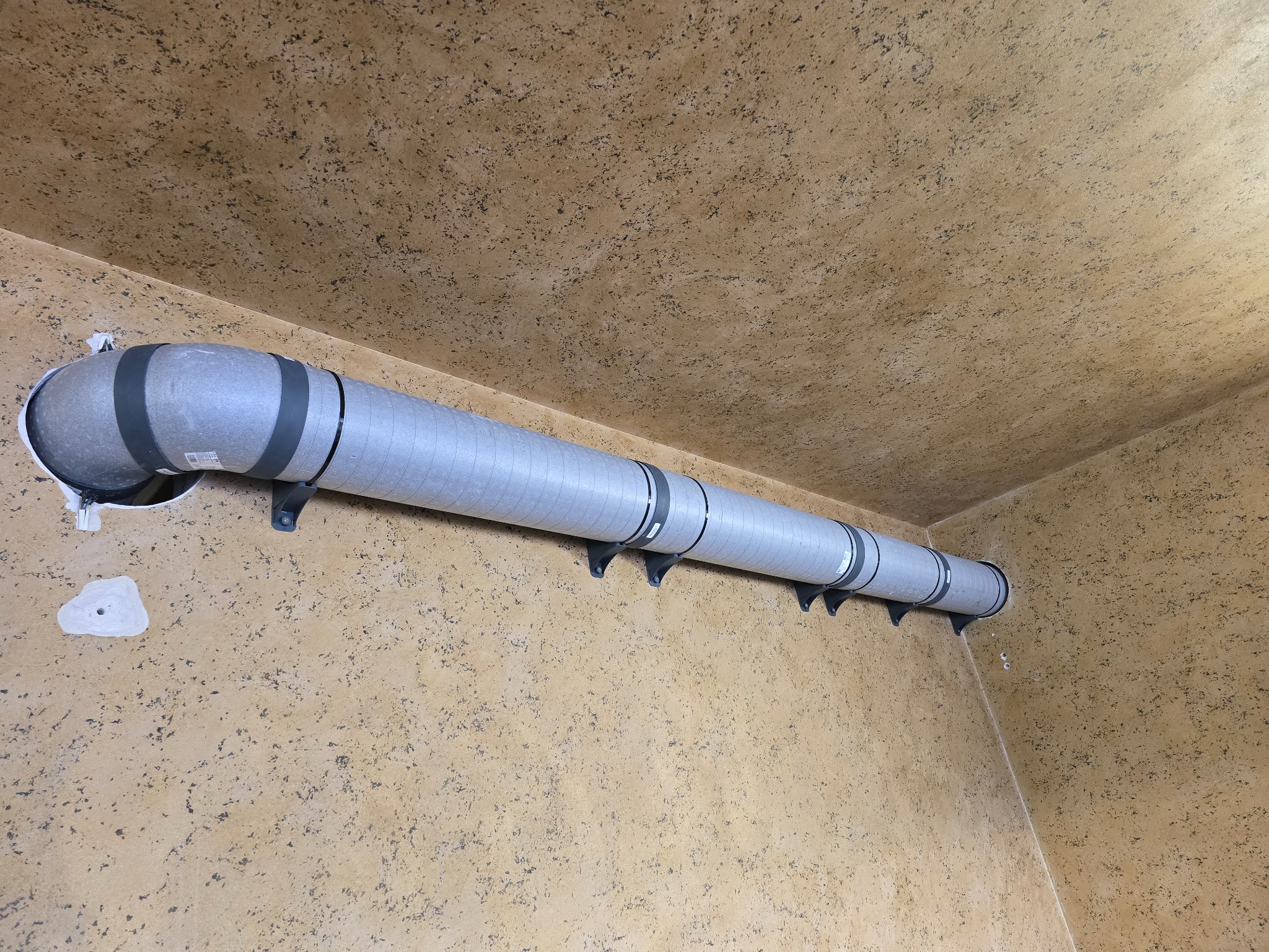 Szary przewód wentylacyjny z czarnymi obręczami, zamocowany do ściany o nierównej, beżowej fakturze, widoczny z perspektywy narożnika pomieszczenia.