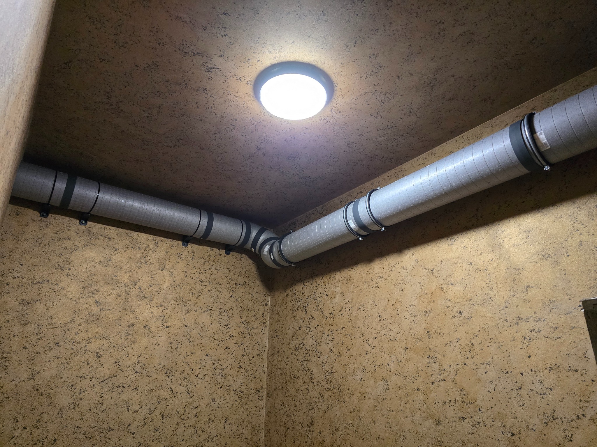 Szare, metalowe rury wentylacyjne z czarnymi obejmami, zamontowane pod sufitem w rogu pomieszczenia o beżowych ścianach. Centralnie umieszczona lampa sufitowa.