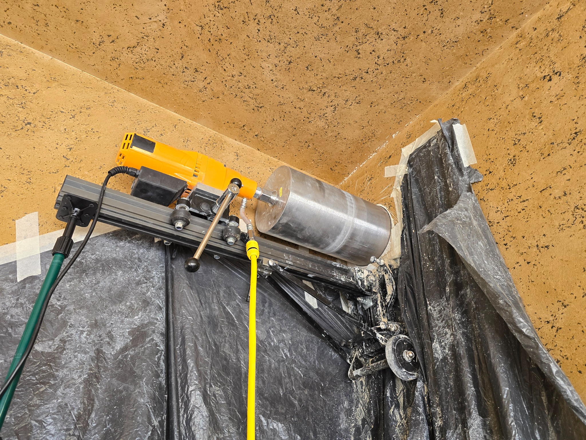 Wiertnica diamentowa w trakcie pracy, mocowana do ściany w rogu pomieszczenia. Widoczne przewody i osłona z folii. Żółto-pomarańczowa wiertarka.