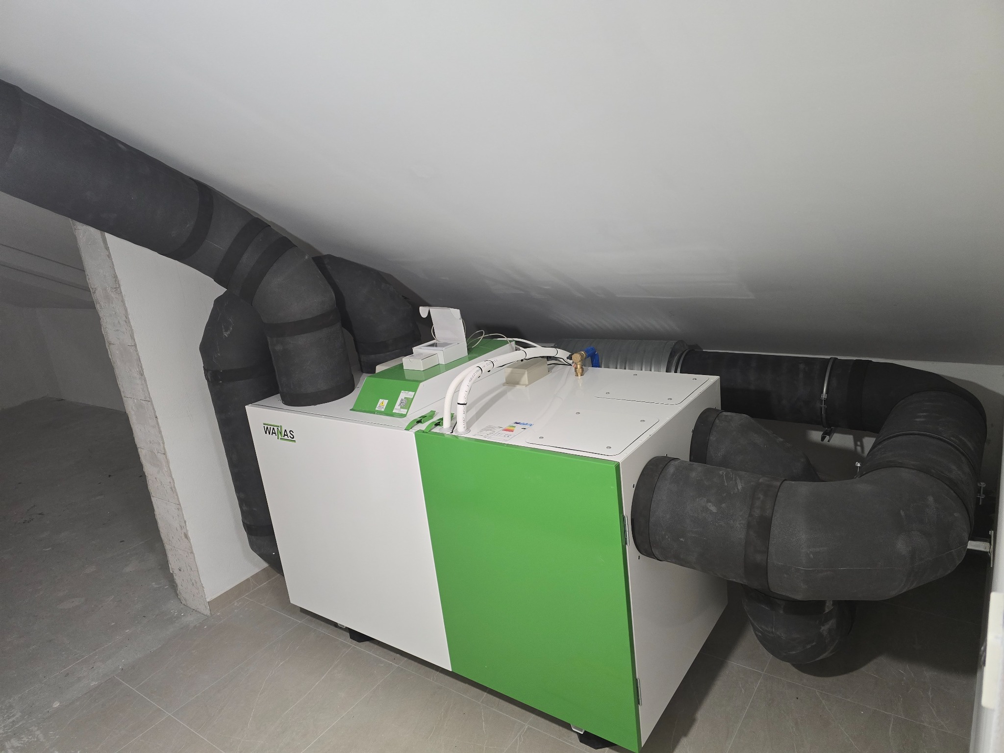 Rekuperator WANAS z podłączonymi przewodami wentylacyjnymi w pomieszczeniu ze skośnym sufitem. Widoczne elementy instalacji i wykończenia.