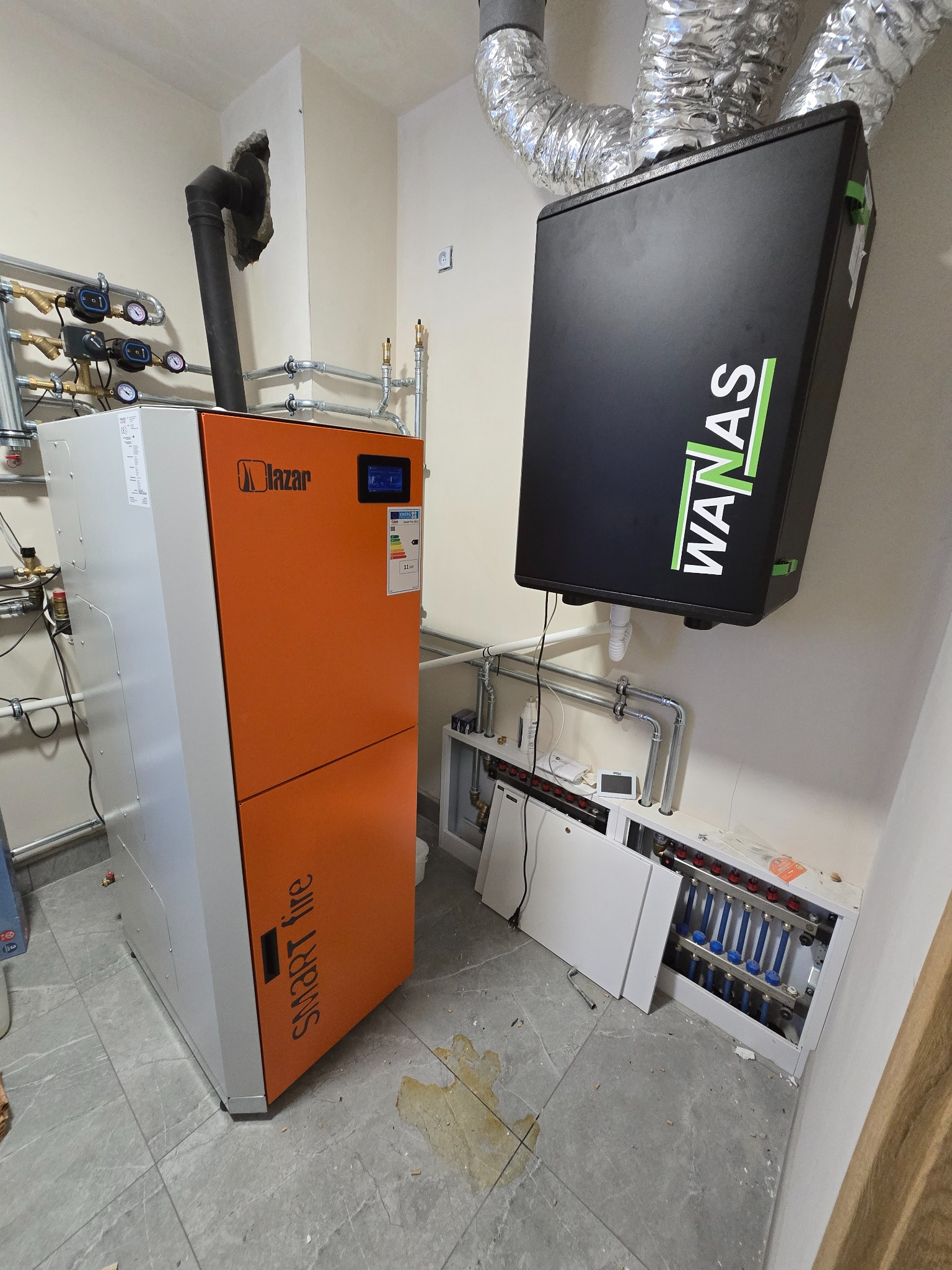 Pomarańczowo-szary piec Lazar Smart Fire z czarnym kominem i czarna pompa ciepła Wanas z instalacją hydrauliczną w pomieszczeniu z szarą podłogą.