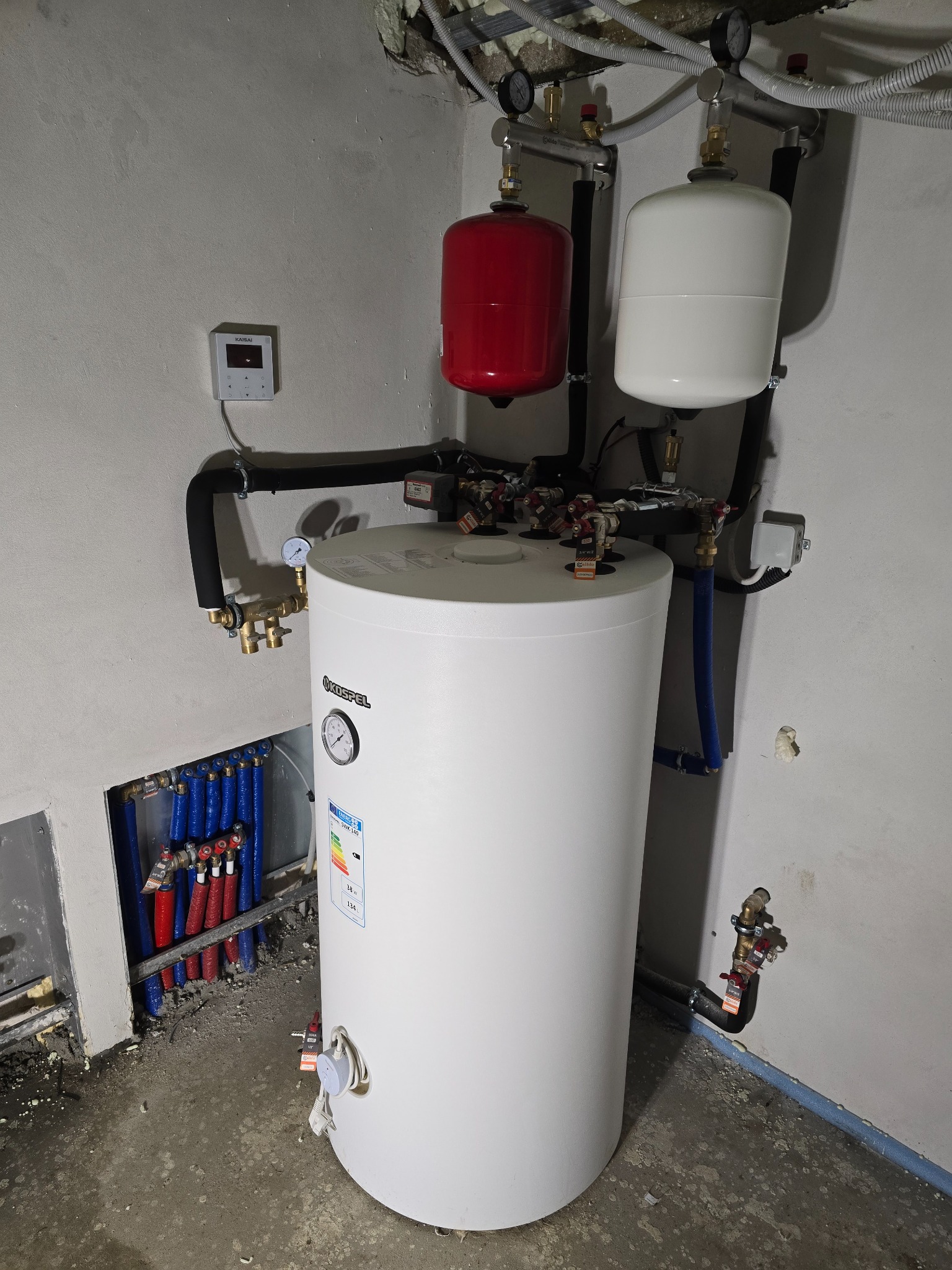 Biały bojler Kospel z manometrem, czerwony i biały zbiornik wyrównawczy, rury instalacji CO w ścianie. Widok na kompletną instalację grzewczą w kotłowni.