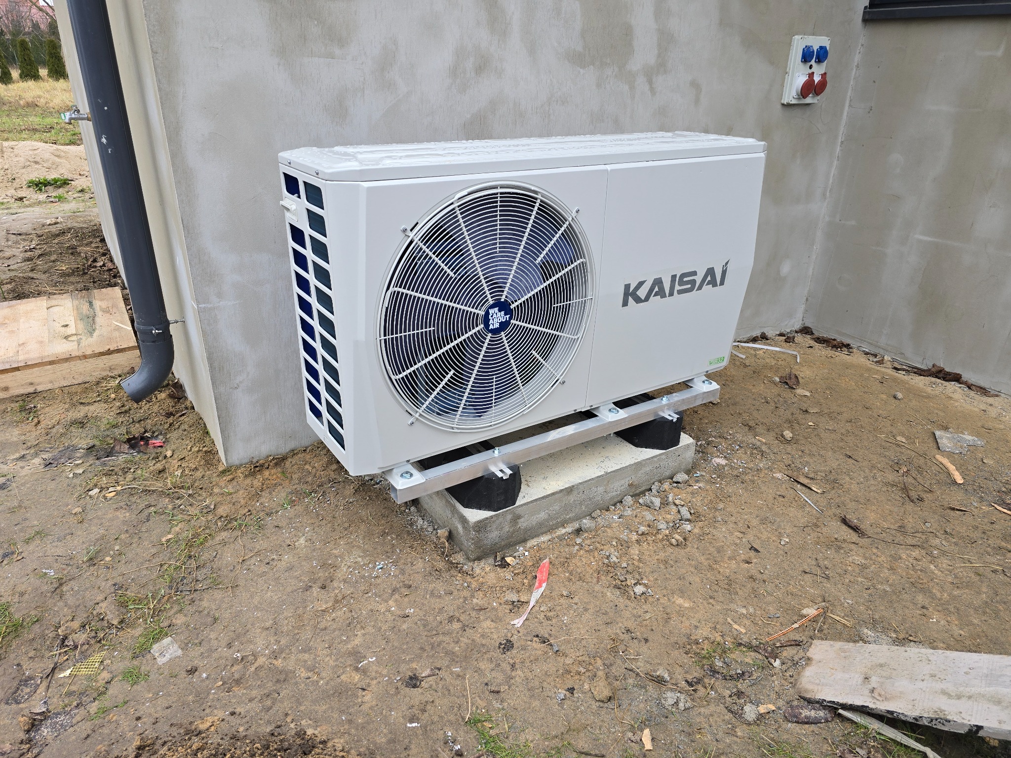 Biała pompa ciepła Kaisai zamontowana na betonowym fundamencie przy ścianie budynku z widocznymi gniazdami elektrycznymi. Widoczny fragment rynny.