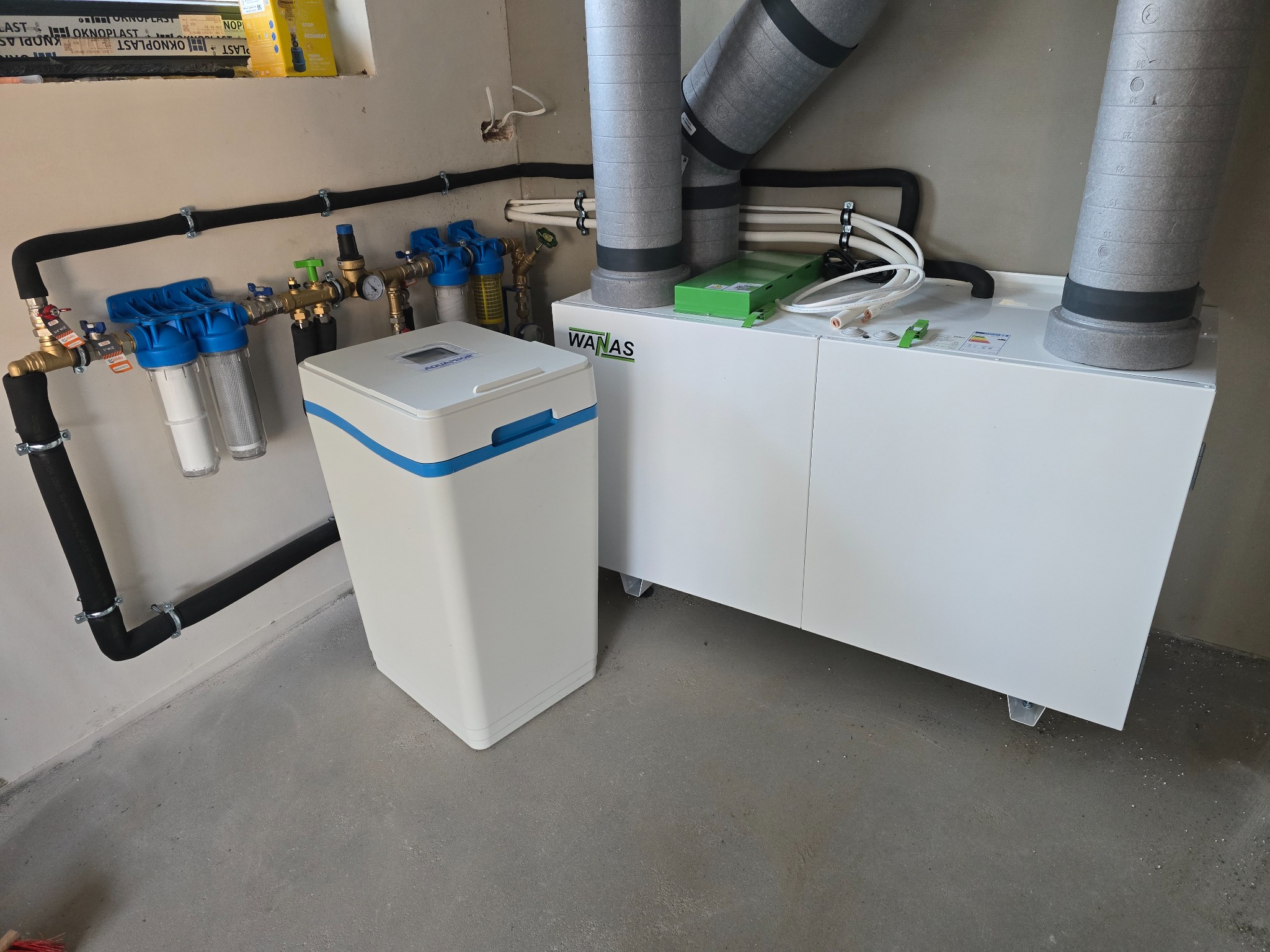 Biały zmiękczacz wody Aquaphor obok rekuperatora WanAs z szarymi przewodami wentylacyjnymi. Instalacja hydrauliczna z filtrami i rurami w izolacji.
