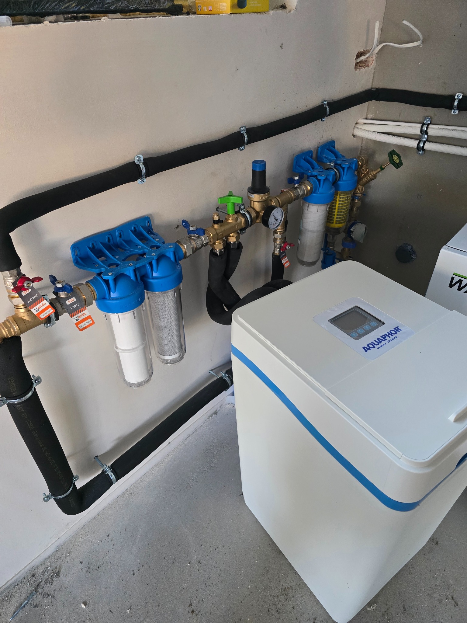 Instalacja filtrów wody Aquaphor z rurami w izolacji, zaworami Calido i manometrem. Widoczne mocowania do ściany. Filtr wody z wyświetlaczem.