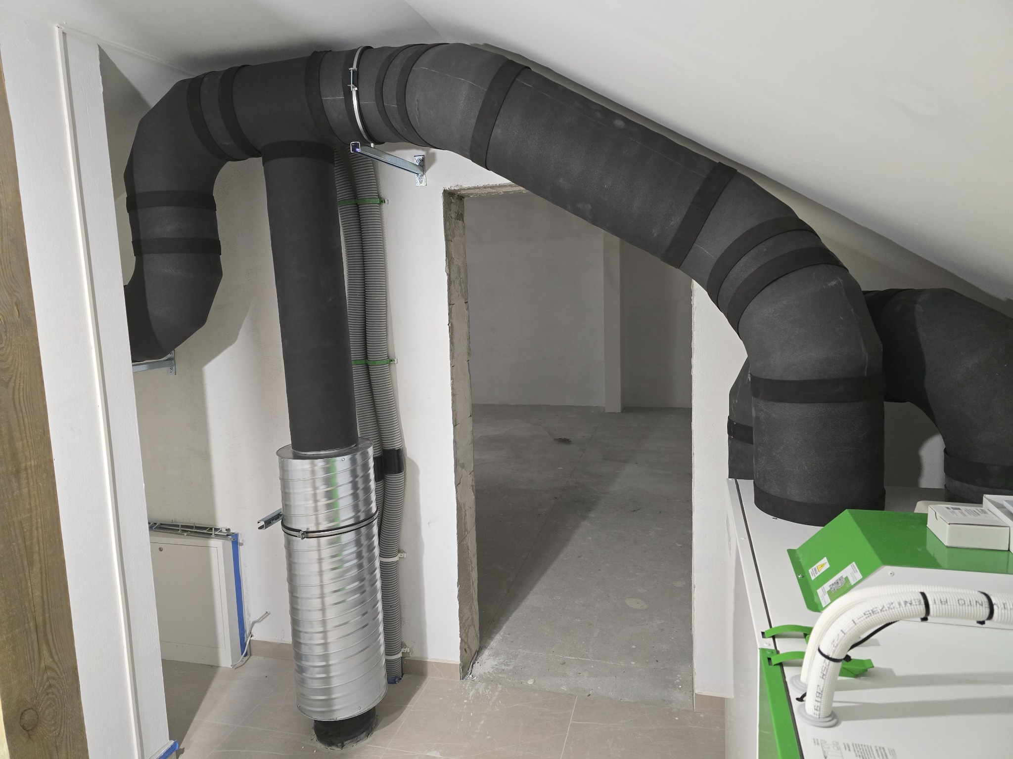 Czarna, izolowana rura wentylacyjna z metalowymi obejmami, zamontowana w jasnym pomieszczeniu z widocznym fragmentem instalacji i otwartym przejściem do innego pokoju.