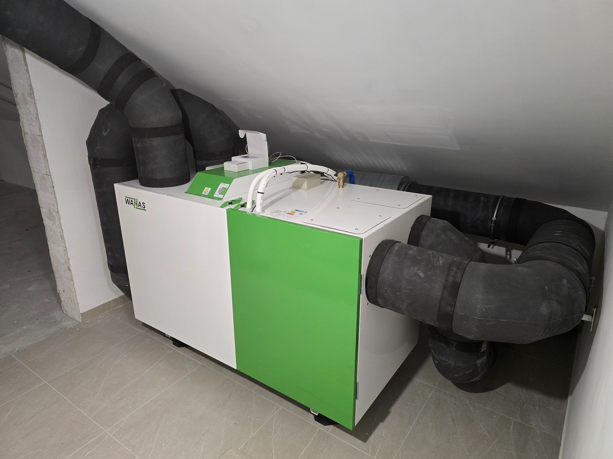 Biało-zielona centrala wentylacyjna z podłączonymi czarnymi przewodami wentylacyjnymi w pomieszczeniu na poddaszu. Widoczne elementy instalacji i logo producenta.