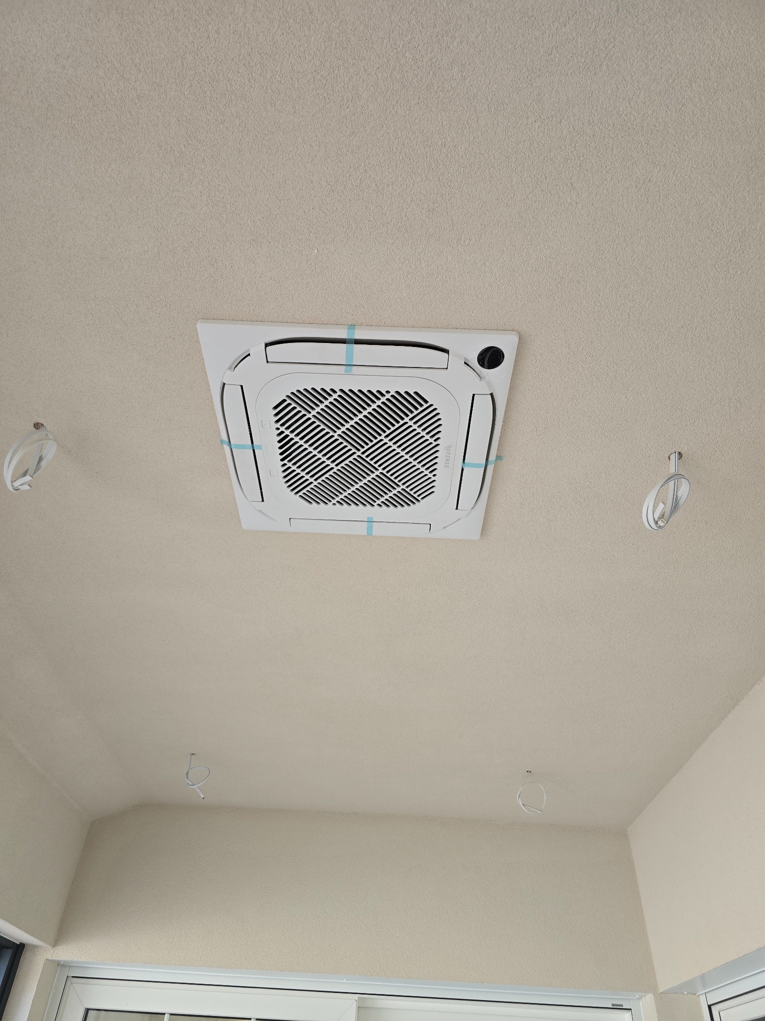 Zamontowana, biała klimatyzacja kasetonowa na suficie z widocznymi przewodami elektrycznymi i taśmą zabezpieczającą. Widok z dołu na jasne wnętrze.
