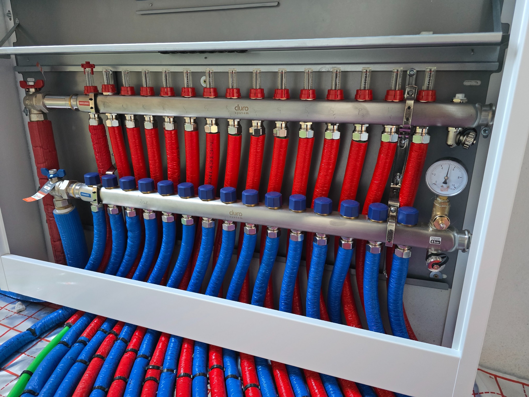 Rozdzielacz ogrzewania podłogowego w szafce, z widocznymi rurami w kolorach czerwonym i niebieskim. Manometr i zawory regulacyjne. System DURO.
