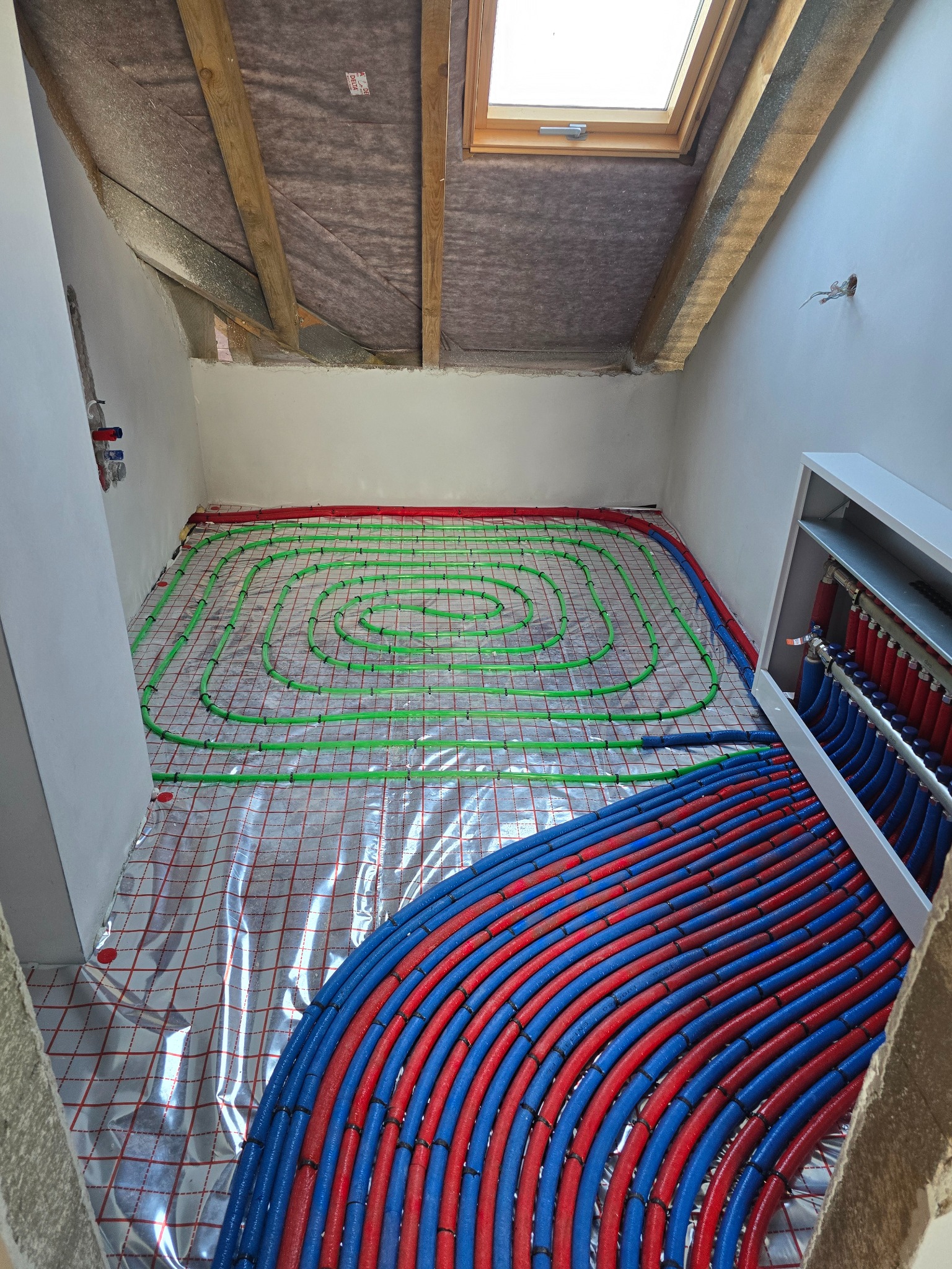 Instalacja ogrzewania podłogowego w poddaszu: zielone i czerwone rury ułożone spiralnie na folii, z rozdzielaczem z niebiesko-czerwonymi rurami. Widoczne okno dachowe.