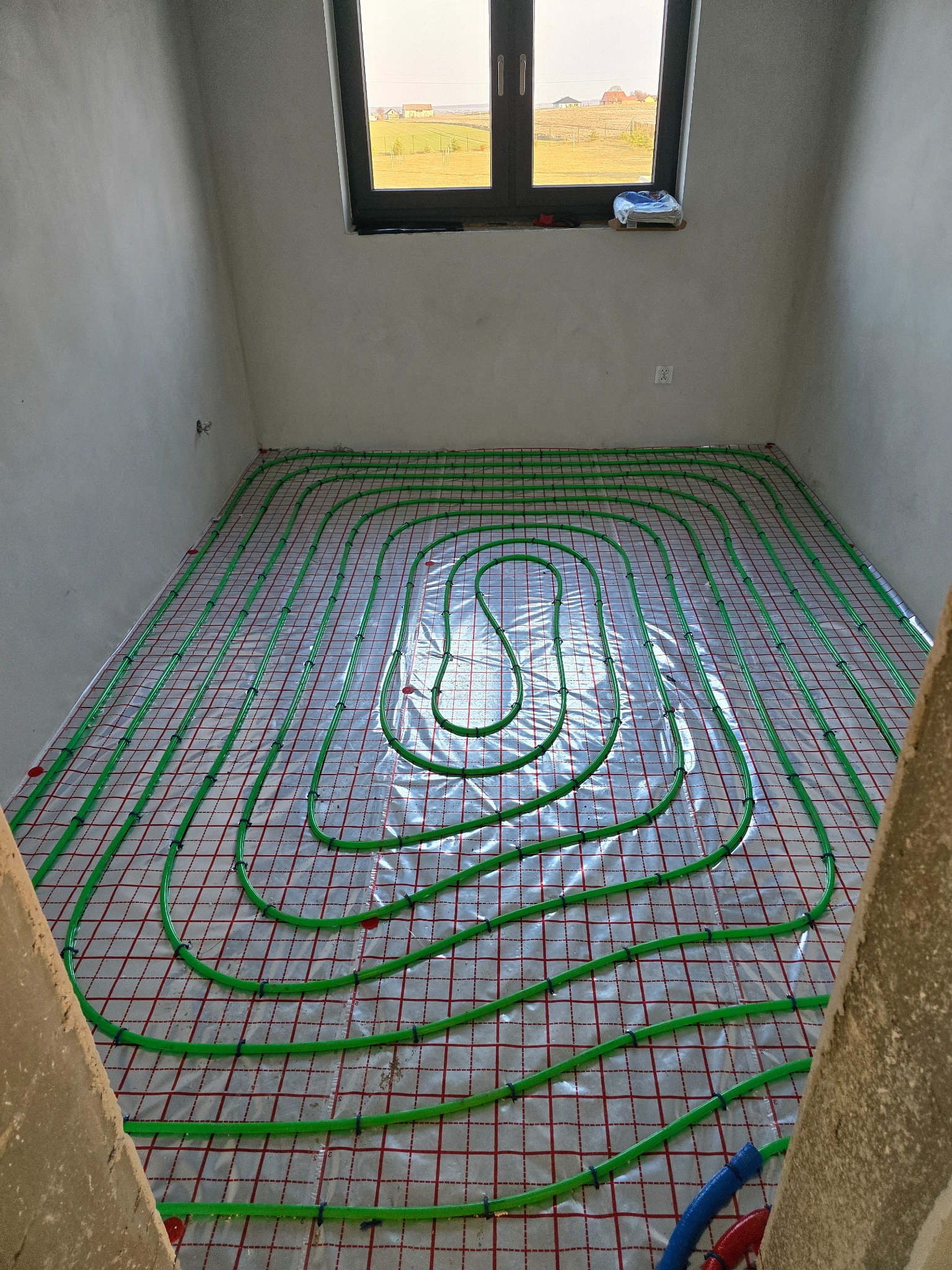 Zielone rury ogrzewania podłogowego ułożone spiralnie na folii z czerwoną kratką w surowym pomieszczeniu z oknem. Widoczne fragmenty nieotynkowanych ścian.