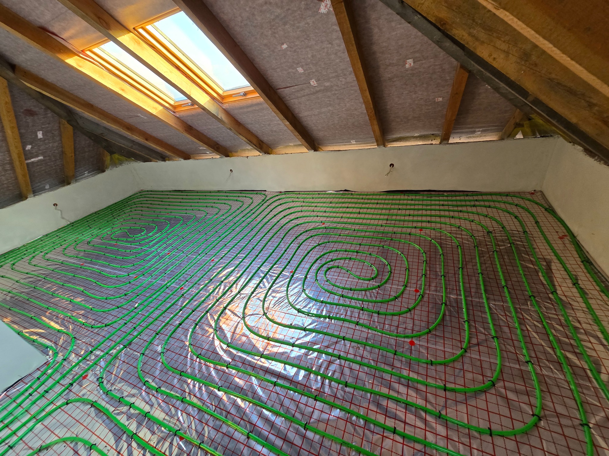Zielone rury ogrzewania podłogowego ułożone spiralnie na folii z czerwoną siatką w pomieszczeniu z oknem dachowym i drewnianą konstrukcją dachu.