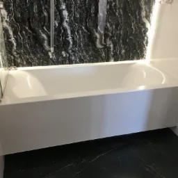 Nowoczesna, biała wanna z parawanem nawannowym na tle ściany wyłożonej czarnym marmurem z białymi żyłami, oświetlona od góry.