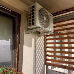 Zewnętrzna jednostka klimatyzacji Samsung zamontowana na elewacji balkonu, widoczne przewody w białej listwie, obok okno z czerwonymi kwiatami i drewniana osłona balkonu.