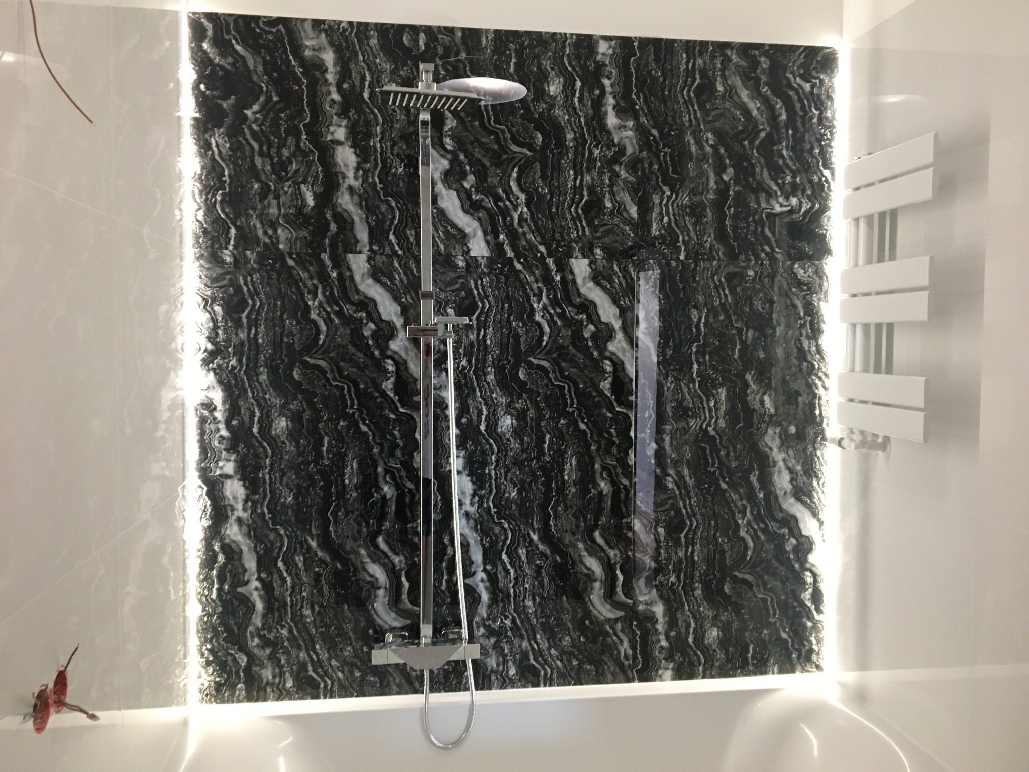 Nowoczesna łazienka z czarną, marmurkową ścianą podświetloną LED, srebrna armatura prysznicowa i biały grzejnik drabinkowy na białej ścianie.