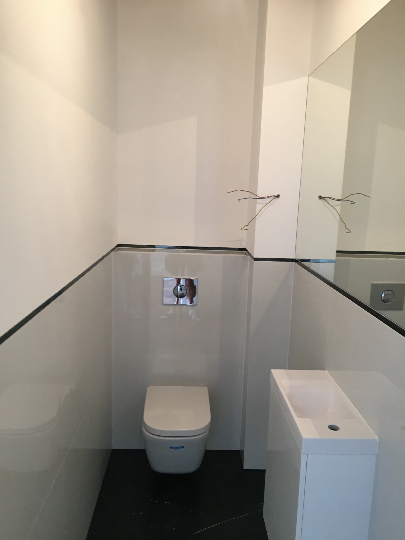 Nowoczesna, minimalistyczna łazienka z białymi płytkami, czarnym akcentem w postaci cienkiego paska i czarnej podłogi, podwieszana toaleta i wąska, biała umywalka z szafką, lustro z widocznymi...