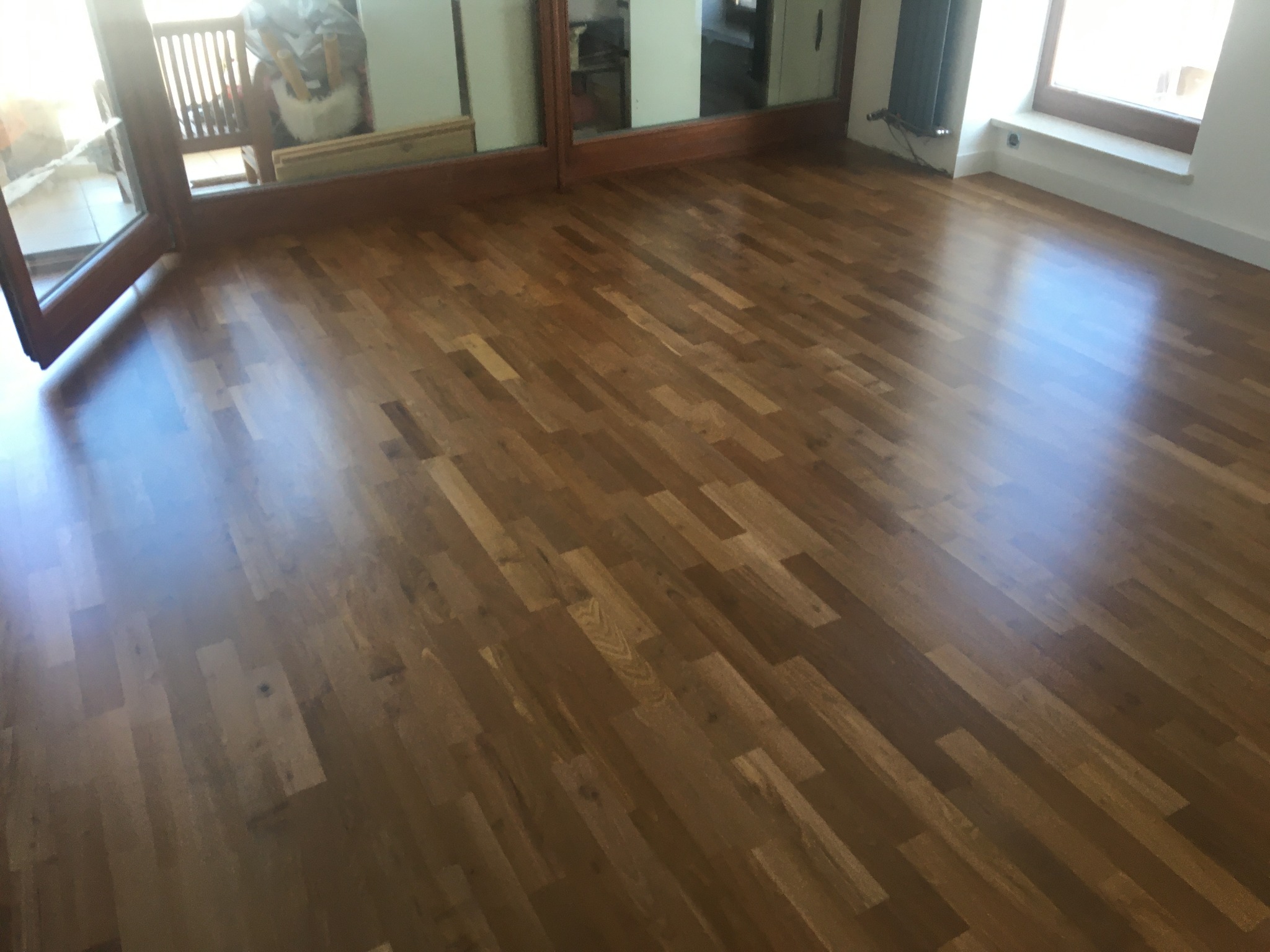 Połyskująca, nowa, drewniana podłoga parkietowa w odcieniach brązu, ułożona w jodełkę, z widocznym fragmentem okna i grzejnika oraz otwartym oknem balkonowym.