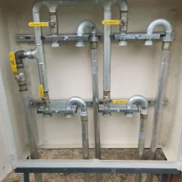 Instalacja gazowa w skrzynce: rury stalowe z żółtymi zaworami, mocowane na szynach. Widoczne połączenia, elementy montażowe i fragment gruntu.