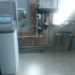 Nowoczesna kotłownia: zmiękczacz wody, miedziane rury instalacji, szare płytki. Schludne wykończenie w neutralnej kolorystyce. Filtr wody z niebieską głowicą.