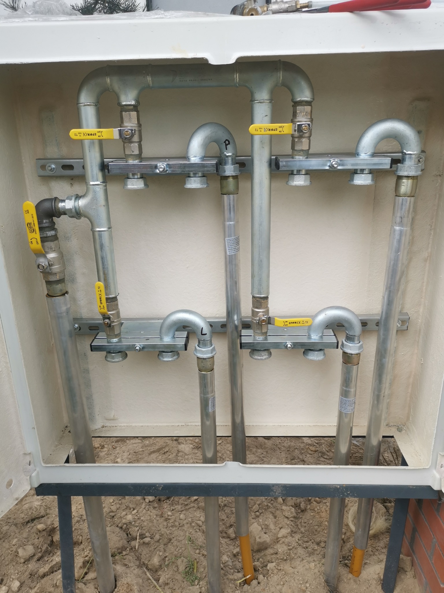 Instalacja gazowa w skrzynce: rury stalowe z żółtymi zaworami, mocowane na szynach. Widoczne połączenia, elementy montażowe i fragment gruntu.