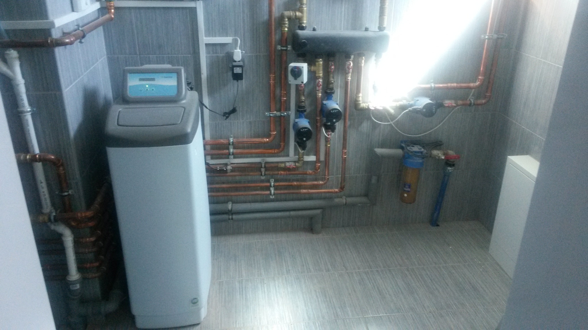 Nowoczesna kotłownia: zmiękczacz wody, miedziane rury instalacji, szare płytki. Schludne wykończenie w neutralnej kolorystyce. Filtr wody z niebieską głowicą.