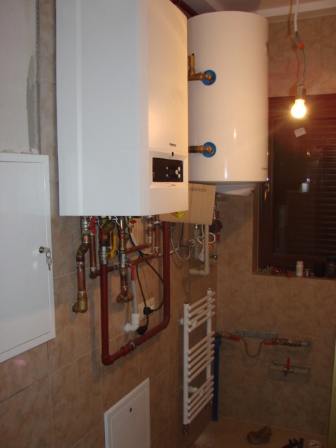 Biały piec gazowy i zasobnik ciepłej wody z widocznymi rurami miedzianymi, zainstalowane w pomieszczeniu wyłożonym beżowymi płytkami. Grzejnik drabinkowy w tle.