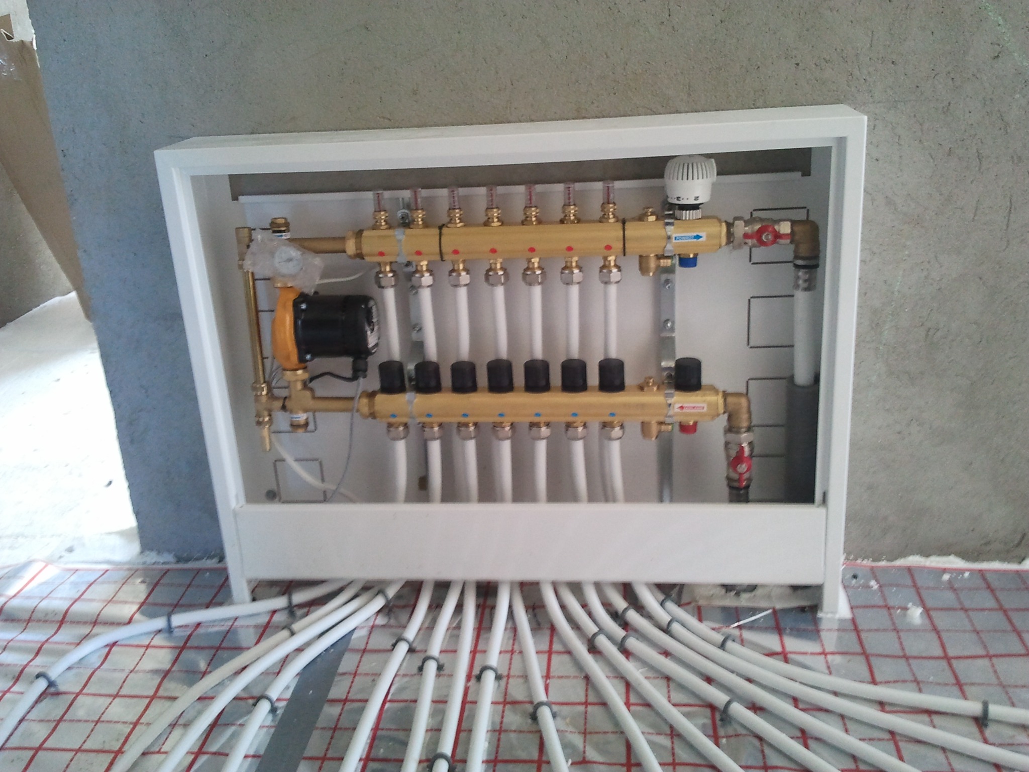 Rozdzielacz ogrzewania podłogowego w białej obudowie, z widocznymi rurami na folii grzewczej, gotowy do zalania posadzką. Precyzyjny montaż.