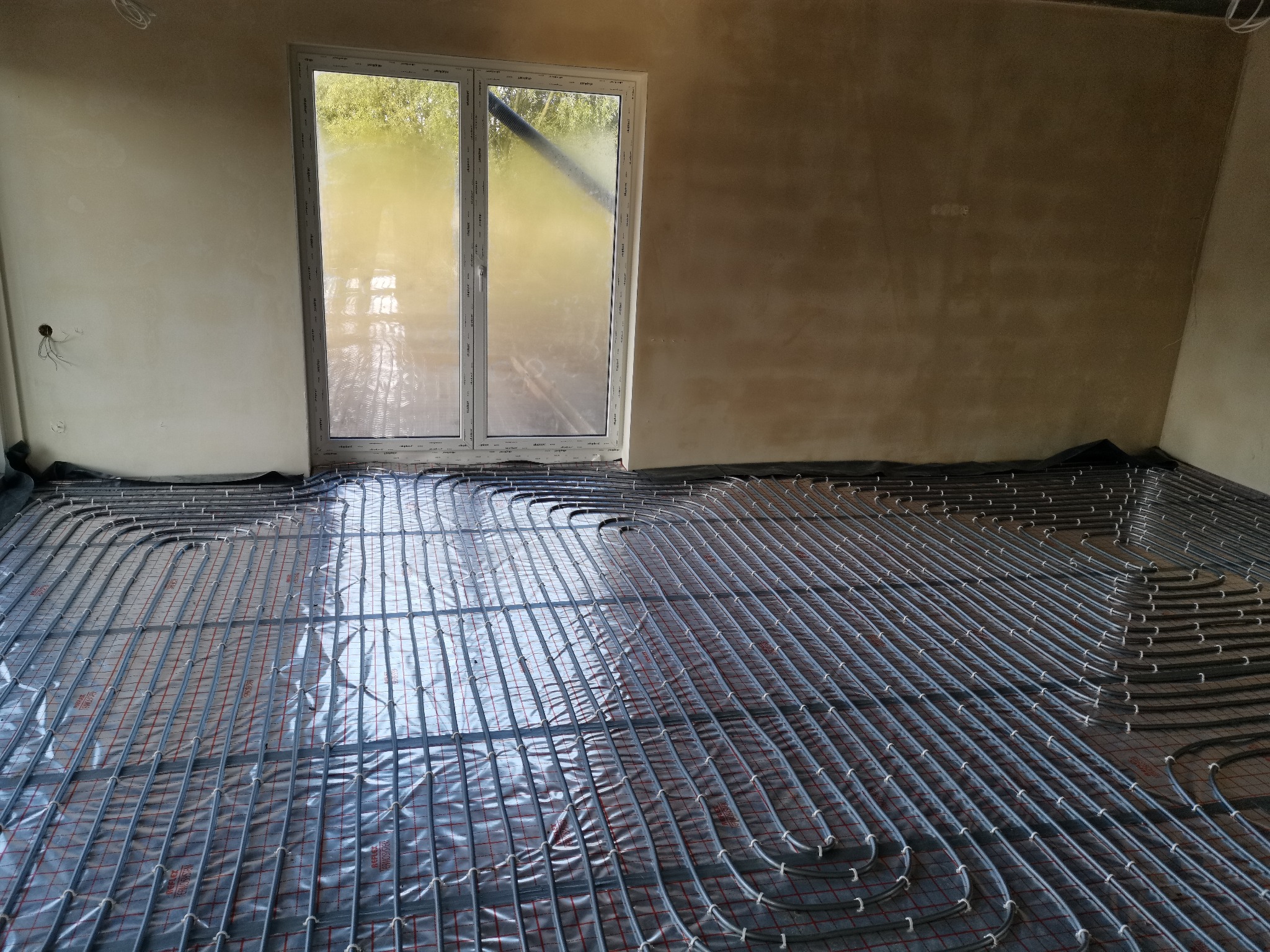 Szare rury ogrzewania podłogowego ułożone na folii z czerwoną kratką w surowym pomieszczeniu z oknem. Widoczne ściany w stanie deweloperskim.