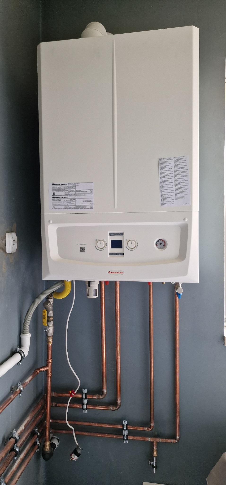 Biały, wiszący piec gazowy Immergas z miedzianymi rurami instalacji grzewczej na szarej ścianie. Widoczne przyłącza i manometr.