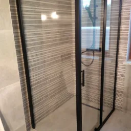 Szklana kabina prysznicowa z czarnymi profilami, zamontowana w łazience z poziomymi, szaro-beżowymi płytkami. Widoczny fragment toalety po lewej stronie.