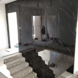 Ściana wyłożona czarnymi, połyskliwymi płytkami imitującymi marmur z białymi żyłami, widoczne surowe schody betonowe i narzędzia glazurnicze na podłodze, odzwierciedlające wnętrze w polerowanej...