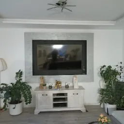 Salon z telewizorem w szarej ramie na ścianie, biała komoda pod spodem z dekoracjami, rośliny doniczkowe po bokach i lampa z abażurem z drewna, sufit z podwieszanym elementem i nowoczesną lampą.