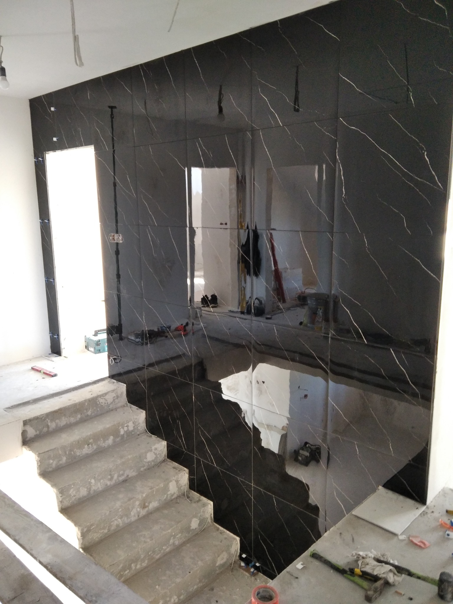 Ściana wyłożona czarnymi, połyskliwymi płytkami imitującymi marmur z białymi żyłami, widoczne surowe schody betonowe i narzędzia glazurnicze na podłodze, odzwierciedlające wnętrze w polerowanej...