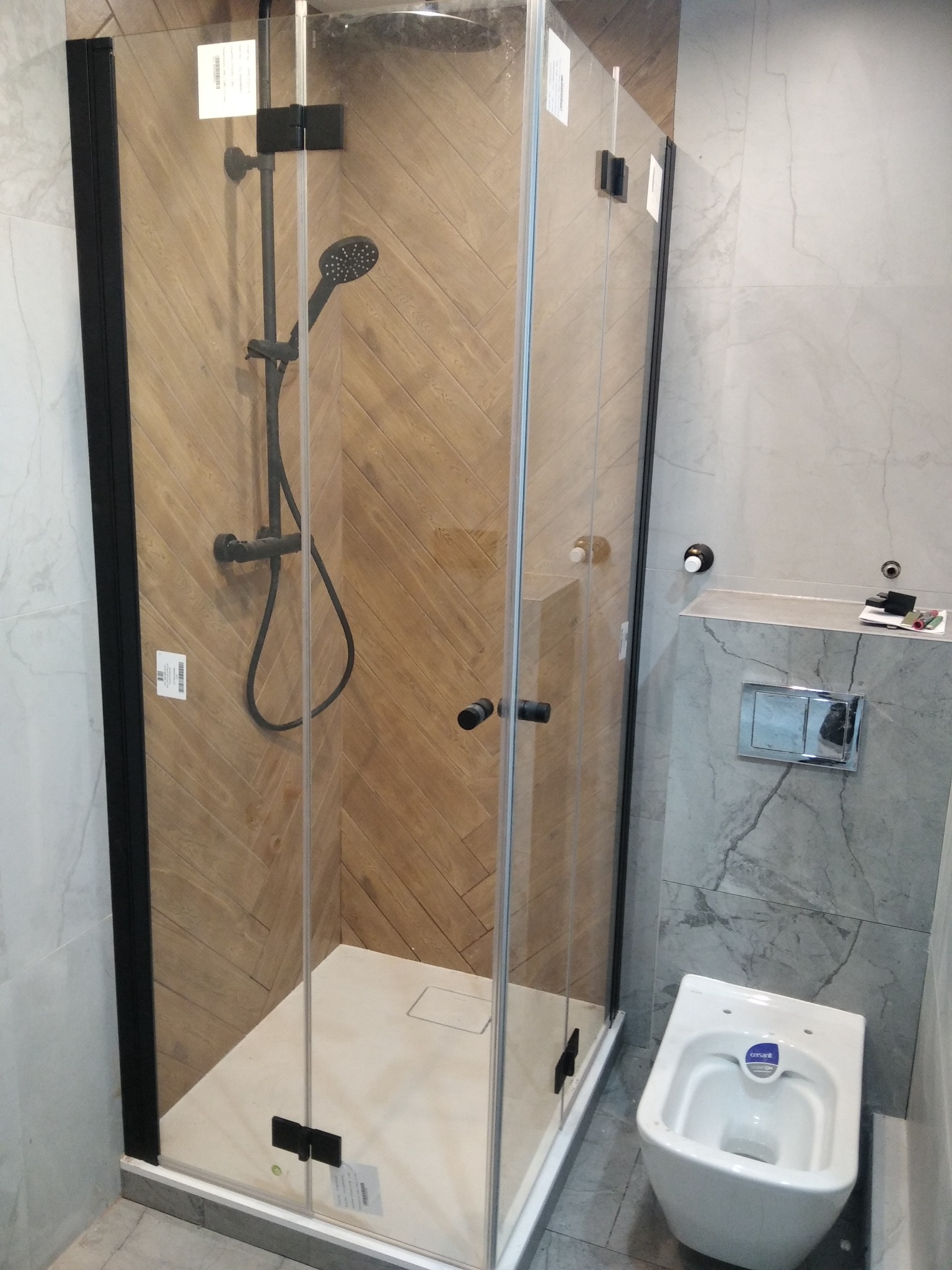 Nowoczesna łazienka z kabiną prysznicową typu walk-in z czarnymi profilami, drewnopodobnymi płytkami ułożonymi w jodełkę wewnątrz kabiny i podwieszaną miską WC.