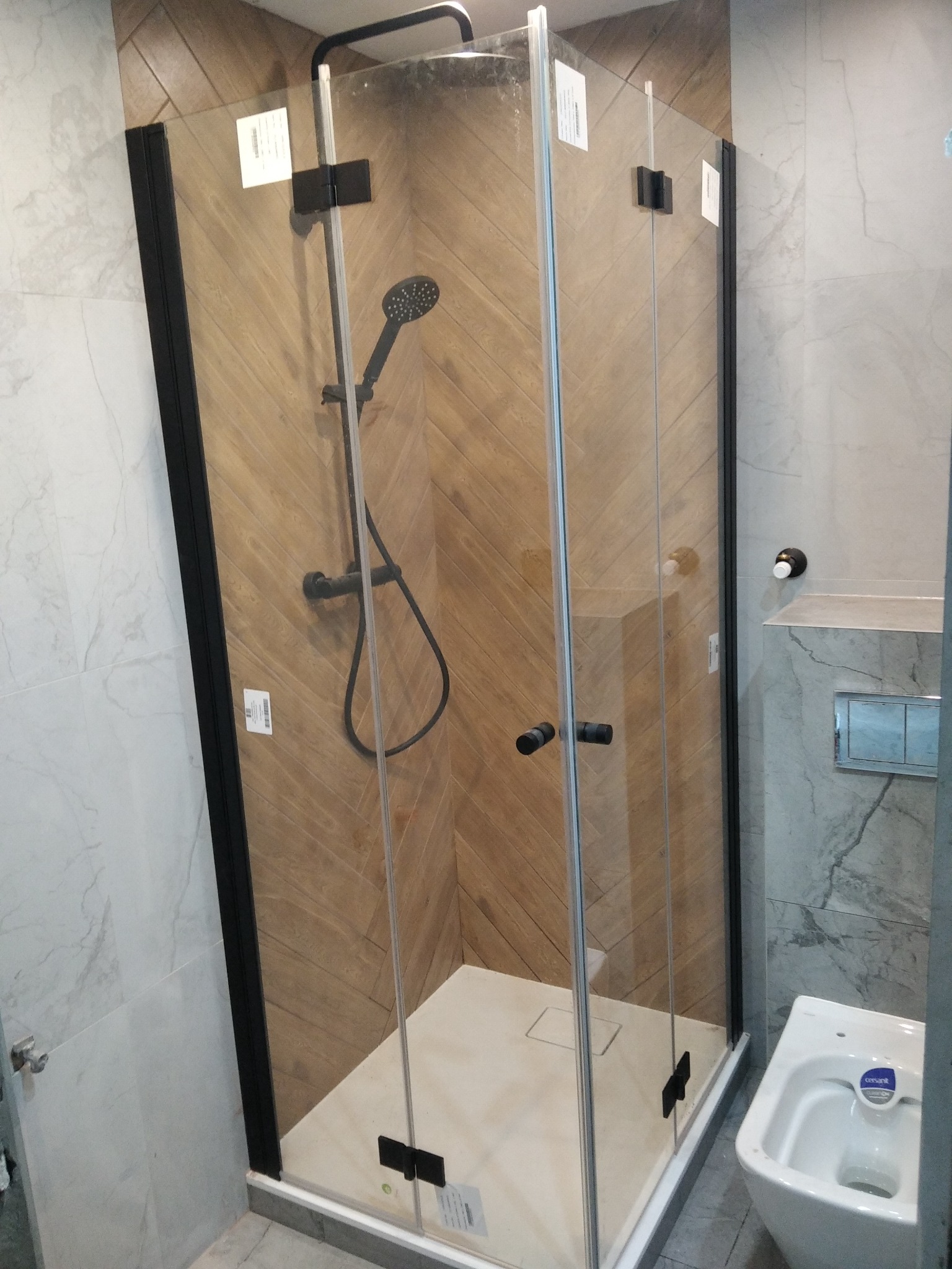 Kabina prysznicowa z czarnymi profilami i armaturą, drewnopodobne płytki ułożone w jodełkę na ścianie, widoczny fragment bidetu.