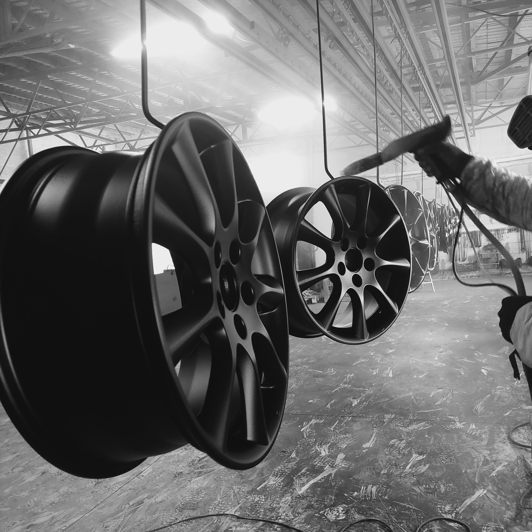 Felgi samochodowe zawieszone w hali, pokrywane czarną farbą proszkową przez pracownika w kombinezonie ochronnym, widoczne z bliska, w tle konstrukcja dachu hali.