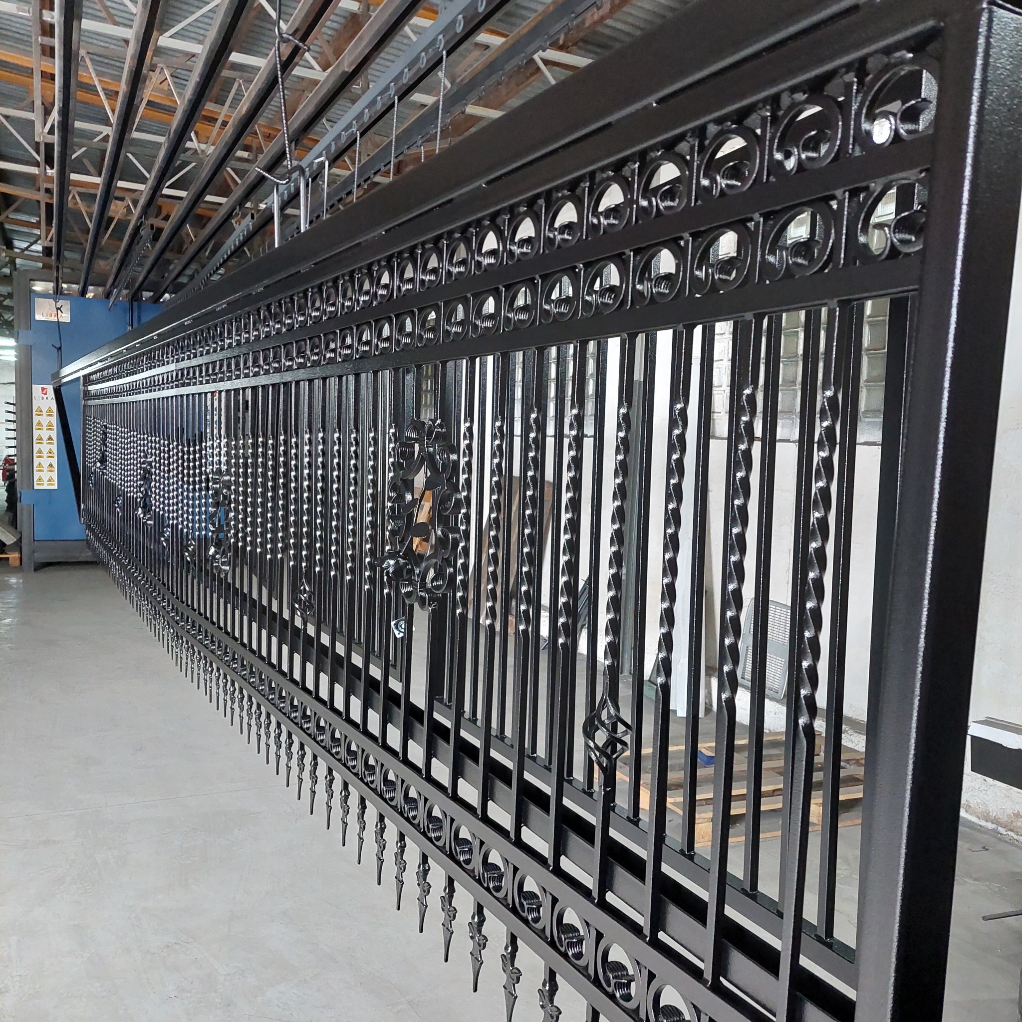 Długa, metalowa brama z ozdobnymi elementami, świeżo po nałożeniu czarnej powłoki, zawieszona na szynach w hali produkcyjnej.