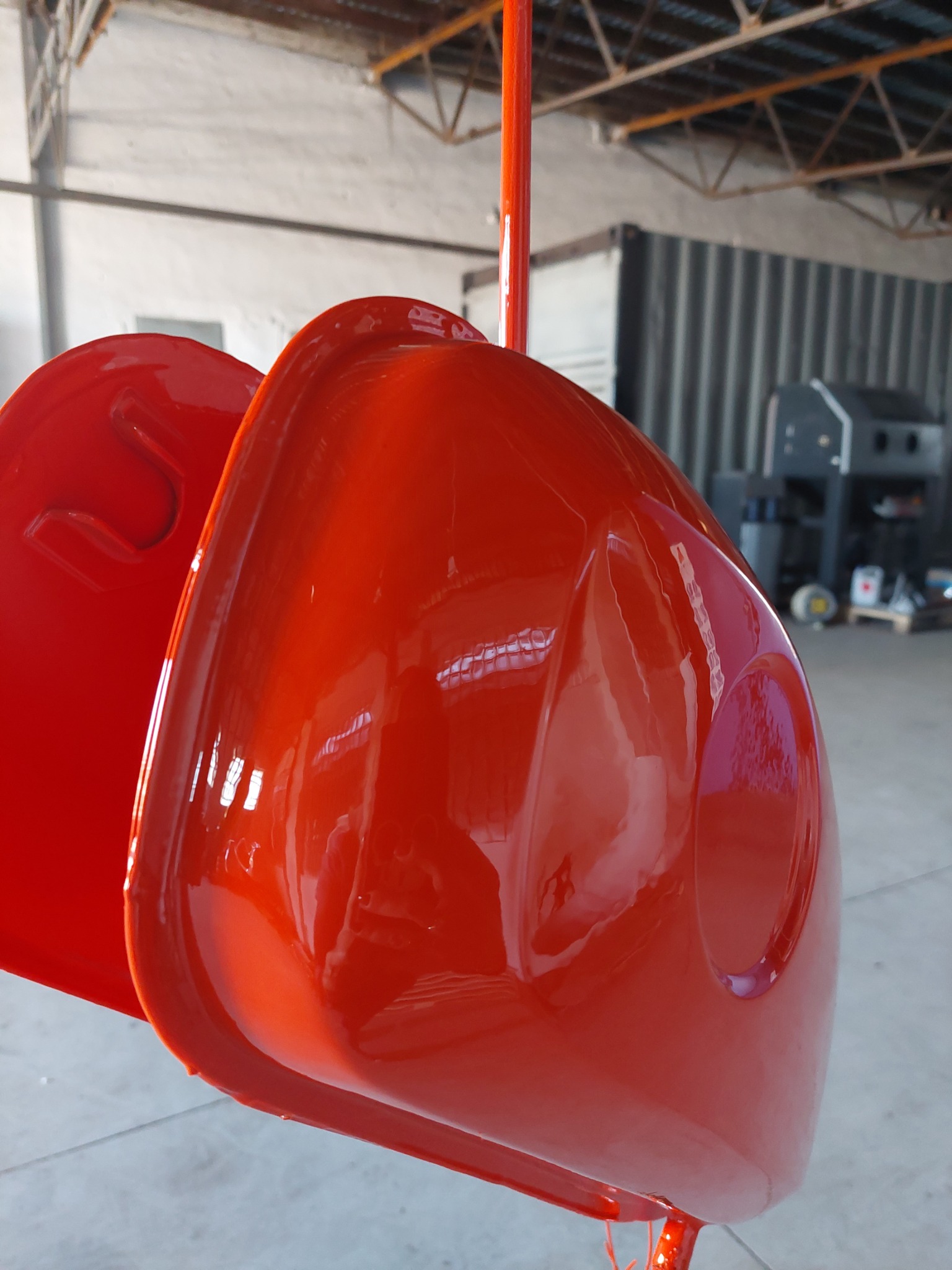 Dwa metalowe elementy, prawdopodobnie części maszyny, zawieszone na pomarańczowym stelażu, świeżo po procesie lakierowania proszkowego na intensywny czerwony kolor, z widocznym połyskiem...