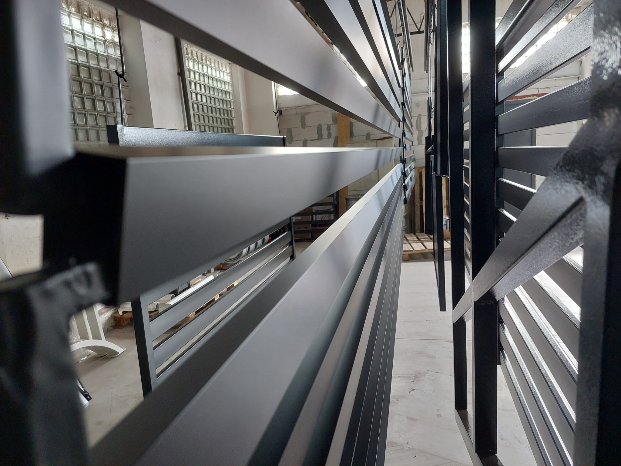 Metalowe elementy konstrukcyjne, prawdopodobnie fragmenty ogrodzenia lub balustrady, zawieszone pionowo na hakach w hali produkcyjnej, po obróbce i pokryciu farbą proszkową, z widocznymi oknami...